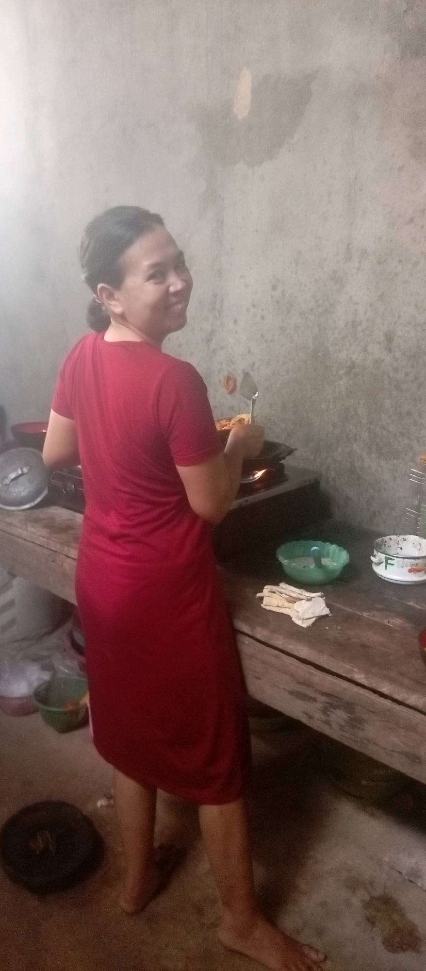 A woman, seen from the back and side, is standing in what appears to be a kitchen in Lubai Ulu, Indonesia. She is wearing a long, plain red dress that reaches her ankles. Her hair is pulled back, and she is smiling as she looks over her left shoulder towards the camera. Her right hand holds a metal spatula, which she is using to stir something in a pan on a gas stove. Flames are visible beneath the pan. The kitchen environment is rustic, with a concrete wall in the background and a wooden table in the foreground. On the table are several items, including a green bowl, a white bowl with a floral pattern, and a stack of folded white cloths. To the left of the stove, a large metal pot is visible, and on the floor to the left of the woman's feet is a black circular object, possibly a weight. The floor is concrete and appears damp in places. The lighting in the scene is dim, and there is some haze or smoke in the air, particularly around the top left of the image.