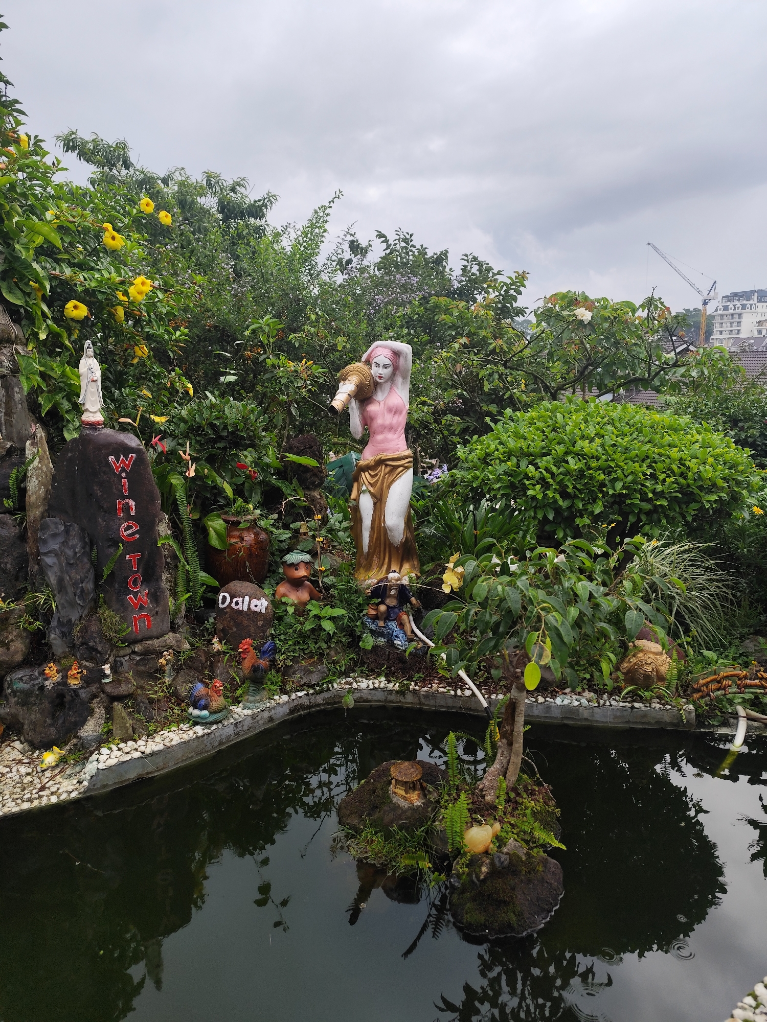 This vertical shot captures a densely decorated garden featuring a pond and an array of statues under an overcast sky. In the foreground, a dark pond reflects the surrounding vegetation, with two small rock islands holding miniature plants, a bonsai-like tree, and a tiny house figurine. The pond's edge is lined with white pebbles and several small, colorful ceramic figures, including two roosters and a fish.
Dominating the midground is a tall statue of a fair-skinned woman with pink hair, draped in a pink top and golden skirt, carrying a large jug on her shoulder. To her left, a white statue, possibly Guanyin, stands atop a dark rock. This rock bears the words "Wine Town" painted vertically in red. Below it, another rock is inscribed with "Dalat" in white letters, confirming the location. Various other small statues, such as a gnome-like figure and several unseen decorative items, are interspersed among the lush green foliage and yellow flowering bushes that fill the garden.
The background reveals more trees, and beyond them, modern buildings and a construction crane are visible against the cloudy, grey sky. The scene is illuminated by the soft, diffused light typical of an overcast day, highlighting the vibrant greens and colorful statues. The overall impression is a whimsical and intricately designed garden, likely a tourist attraction or part of a larger resort in Da Lat, Vietnam.