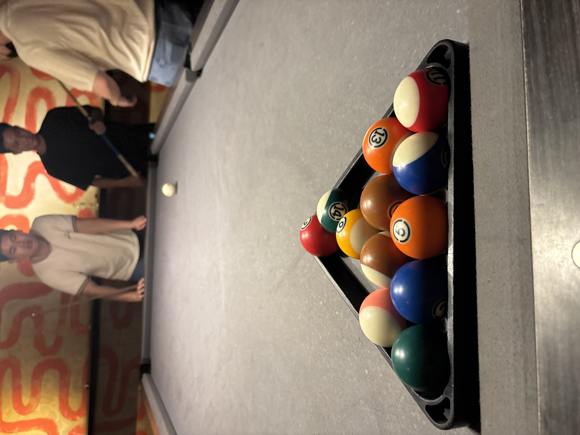 The image depicts a group of young men playing pool in an indoor establishment, likely a bar or entertainment venue. The setting features a pool table with a gray felt surface and a black rack holding a triangle of billiard balls. The balls are predominantly solids and stripes, with visible numbers such as 5, 13, 14, and possibly others. In the background, a wall is decorated with an orange, abstract, wavy pattern.
Three individuals are partially visible. On the left, a young man in a white t-shirt leans over the table. In the center, another young man in a black t-shirt holds a pool cue, appearing to be in the process of playing. On the right, a third individual in a light-colored t-shirt is also visible, facing away from the camera. The overall atmosphere suggests a casual social gathering centered around the game of pool. The specific location in the city of Đà Nẵng, Vietnam, cannot be determined from the visual information provided in the image.