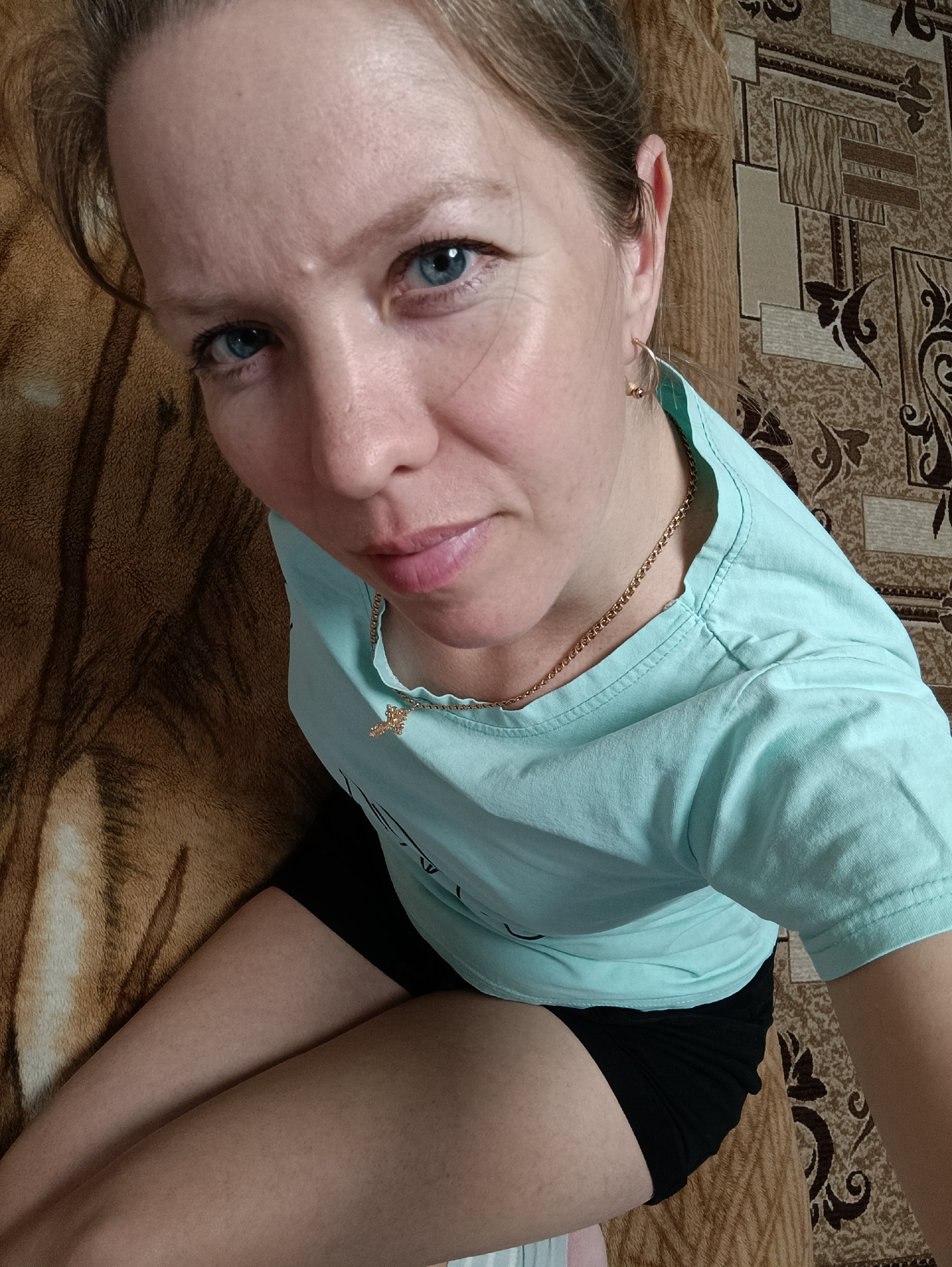 A woman with light-colored hair and striking blue eyes is depicted in a high-angle portrait, looking directly at the camera. She is indoors, likely in a home in Chaikovsky, Russia, seated or reclining against a backdrop of patterned textiles. To her left is a brown and tan patterned blanket or bedding, while to her right, a brown and beige patterned carpet or rug is visible.
She wears a light blue short-sleeved t-shirt, which has some faint, indistinguishable text or design on it, and black shorts. A thin gold chain with a small cross pendant adorns her neck, and she has small gold hoop earrings in her ears. Her expression is calm and direct, with a subtle hint of a smile. The lighting is soft and even, typical of an indoor environment. No specific activity beyond posing for the photograph is evident.