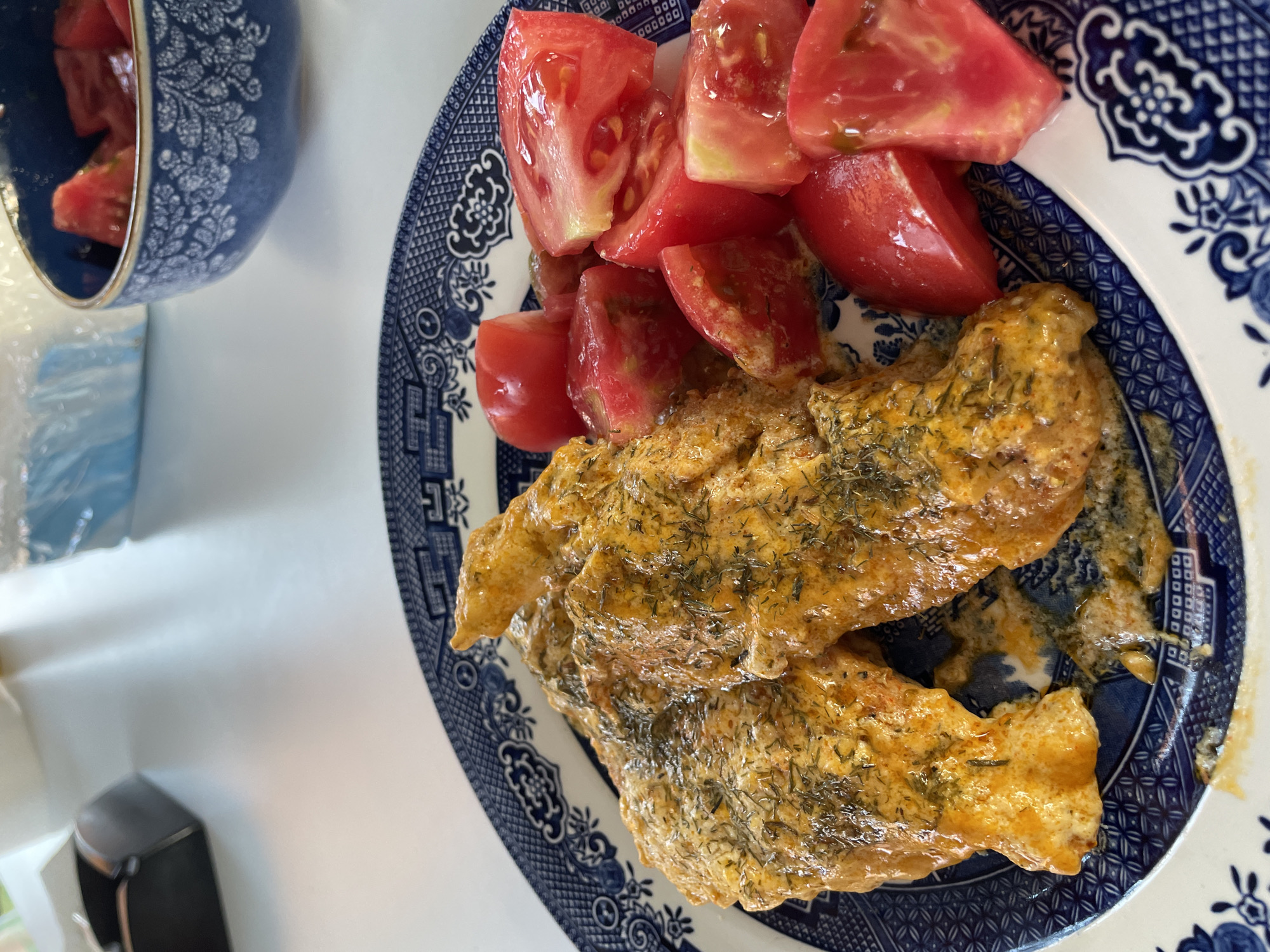 This is a top-down, close-up photograph of a meal served on a plate. The main subjects are two pieces of cooked fish, coated in a creamy, orange sauce and sprinkled with chopped green herbs, likely dill. The fish is presented on a blue and white patterned plate, in the style of Willow pattern. To the left of the fish, on the same plate, are several wedges of ripe, red tomatoes, some of which appear to be cut from cherry tomatoes.
In the background, to the left, is a blue and white patterned bowl containing more tomato wedges. The setting appears to be a table, with a white surface visible to the left of the plate. A partially visible black object, possibly a small appliance or container, is also in the lower left corner of the frame.
The lighting suggests it is daytime. There are no people visible in the image. The meal appears to be freshly prepared and ready to be eaten. The overall impression is of a simple, homemade meal. No visible text is present. The location context is Skala Avdiron, Greece.