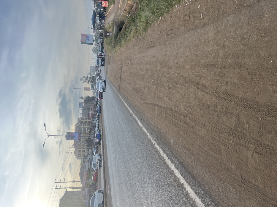 A photograph shows a slightly tilted view of a road with moderate traffic. The road is damp and shiny, with a white line dividing it from a dirt shoulder. Buildings and billboards line the left side of the road. Tire tracks are visible on the dirt shoulder. Overhead power lines stretch toward the sky. The sky is cloudy with hints of blue.
