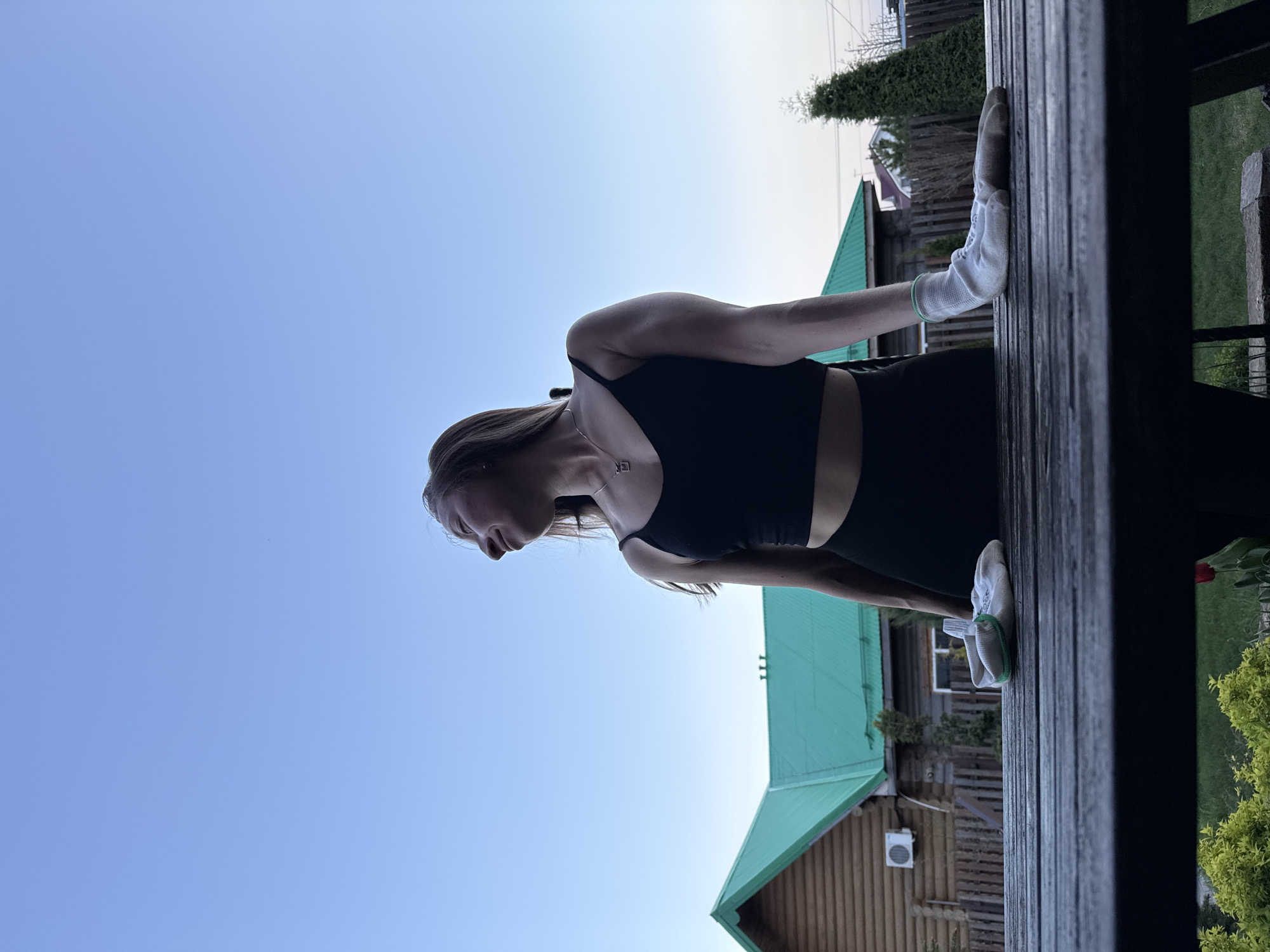 A silhouetted woman is depicted outdoors, likely in Прикамский, Russia, based on the location information. The sky, a gradient from light blue to light gray, is visible. The woman wears a black top and bottoms, and white gardening gloves. She appears to be performing stretching or gardening activities near a wooden structure, possibly a deck. A building with a green roof is in the background.
