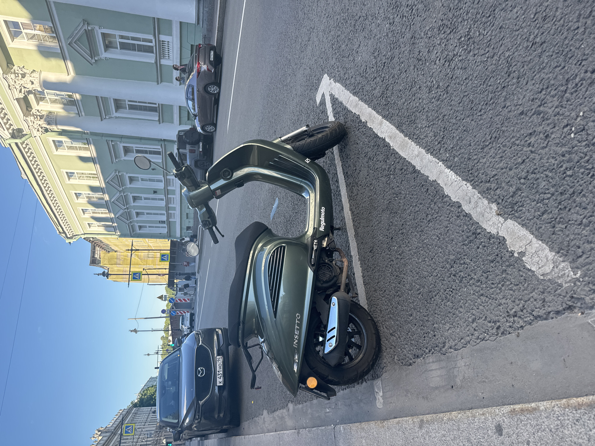 A dark green electric scooter is parked on the side of a street in Saint Petersburg, Russia, with the right rear wheel positioned within a white arrow painted on the asphalt, indicating a directional lane. The scooter has "INSETTO" and "Regulmoto" branding. In the background, a dark gray Mazda CX-5 is parked, and further down the street, a light brown sedan is visible. An ornate, light green building with classical architecture, featuring columns and decorative cornices, lines the right side of the street. Pedestrians are present on the sidewalk in front of the building. The sky is clear and blue, suggesting a sunny day.