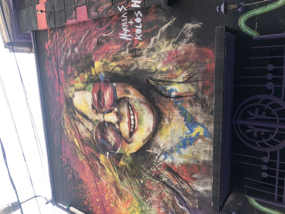 A vibrant mural depicts a person with red-tinted glasses, painted in a loose, expressive style. The background bursts with multicolored streaks and splatters. Adjacent to the mural, a decorative wrought-iron gate painted in purple features musical note motifs. The artwork and gate are mounted on a dark surface with architectural details above. The piece appears to be outdoor public art.