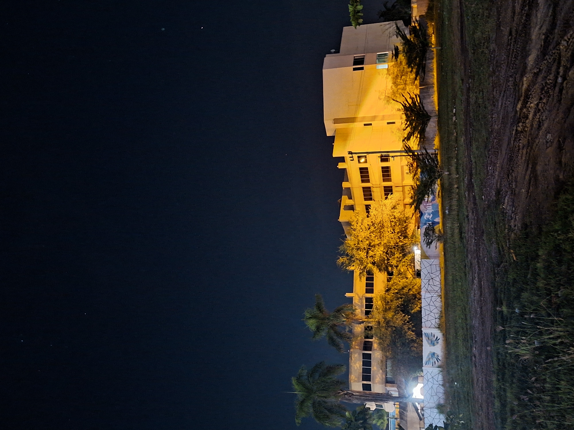 The image depicts a building in Lhokseumawe, Indonesia at night. The building is illuminated with yellow lights. To the right of the building is a grassy area. The sky is dark. There are palm trees in front of the building. A low white wall is visible along the edge of the grass.
