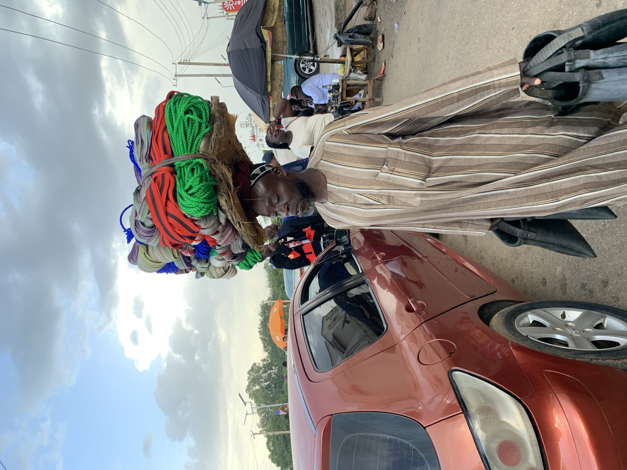 A man in Jos, Nigeria, carries a large stack of colorful ropes and woven items on his head. He wears a striped brown and white tunic and carries two black bags in his hands. He is walking past a red car on a dirt road. In the background, there are people sitting under an umbrella, and a few more cars and trees. The sky is cloudy with patches of blue.