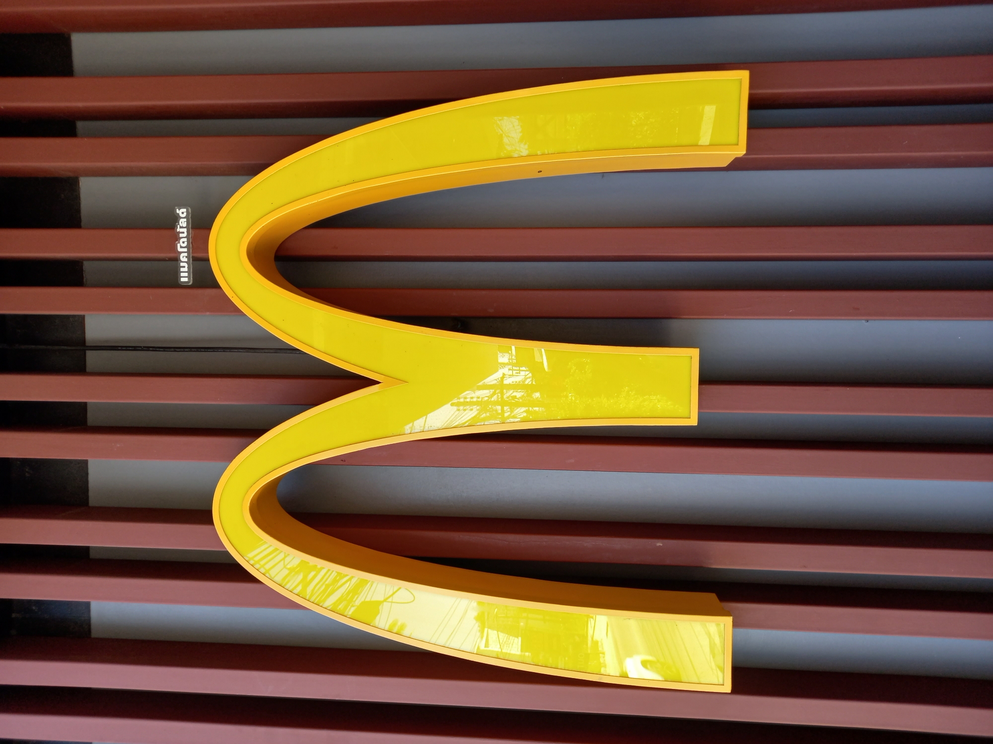 The image shows a close-up view of a large, yellow, three-arched McDonald's logo, also known as the Golden Arches. The logo is illuminated and appears to be made of a glossy material, reflecting the surroundings. It is mounted against a background of horizontal, reddish-brown slats, with some gray sections visible between them.
There are no people or animals in the image. The focus is entirely on the McDonald's logo and the background structure. The lighting suggests it is daytime, as the logo is well-lit and there are reflections indicating natural light. The background slats might be part of a building's facade or an outdoor covering.
There is a small section of text visible on the left side of the image, within the first arch of the logo. The text appears to be in Thai script and reads "แมคโดนัลด์" which translates to "McDonald's".
The overall impression is of a clean and modern signage, likely at a McDonald's restaurant. The location context provided is Baan Bo Phut, Thailand, which would explain the Thai script on the sign. The image is taken from a slightly upward angle, looking at the logo from below.