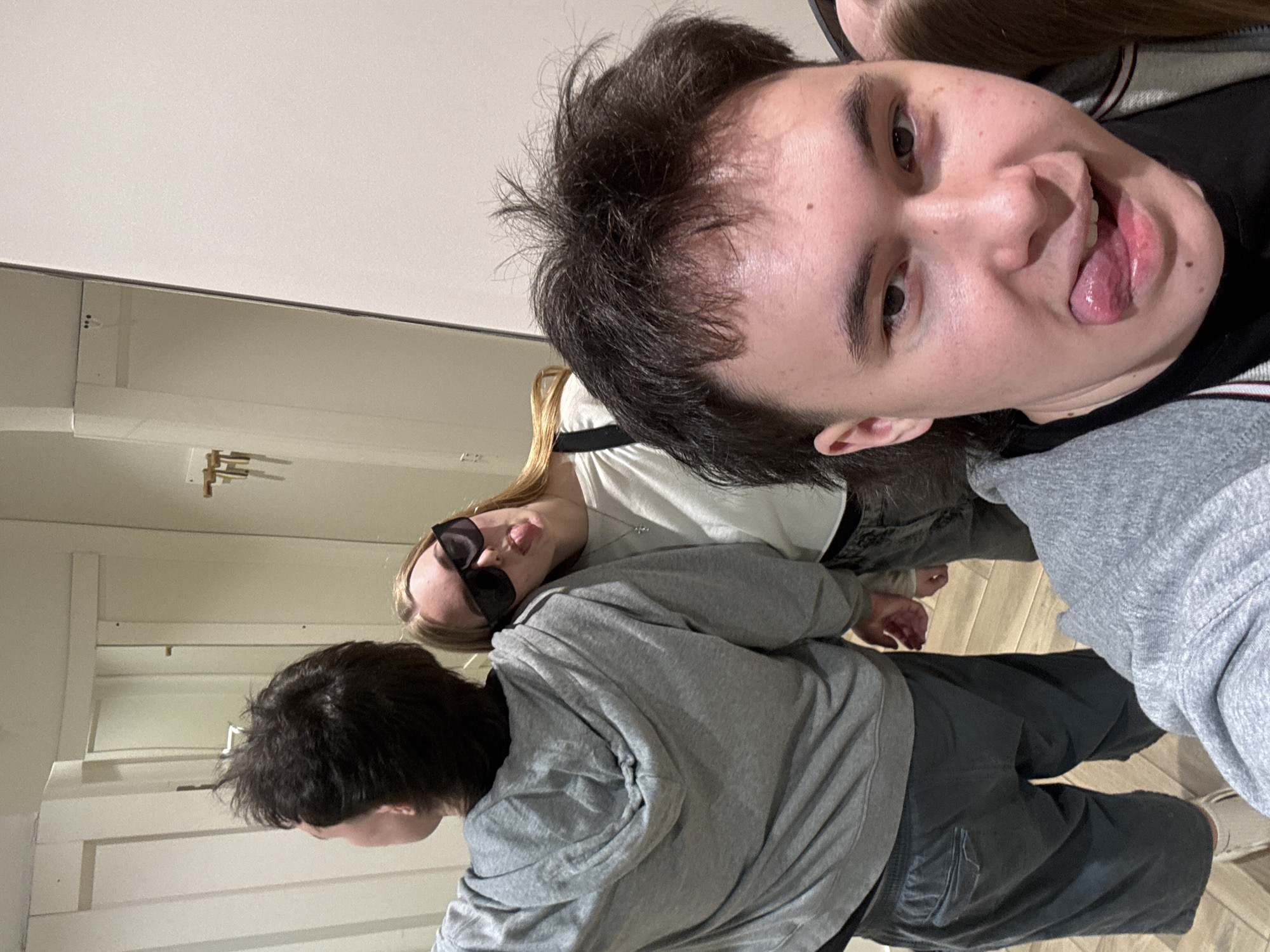 Three individuals are visible indoors. One person, facing the camera directly, is sticking their tongue out. Another individual is wearing sunglasses. A third individual is partially visible with dark hair. All are wearing casual clothing. The background includes a wall and wooden flooring. There is no clear indication of the location being in Perm, Russia.