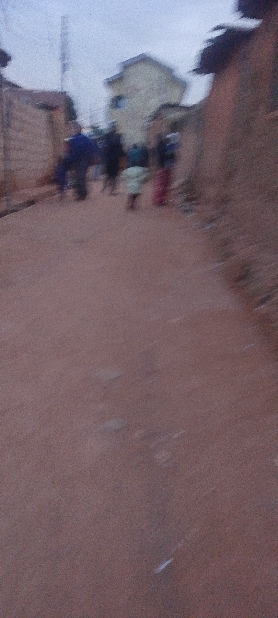 The image depicts a blurry street scene in Gura Top, Nigeria, characterized by a reddish-brown dirt road. Several figures, their details indistinct due to motion blur, are walking along the road. To the left, a building with a brick wall is visible, and further back, a lighter-colored building with a sloping roof stands out. A tall structure, possibly a telecommunications tower, is also present in the background. The sky appears overcast. The overall impression is one of a community street scene with people engaged in everyday movement.