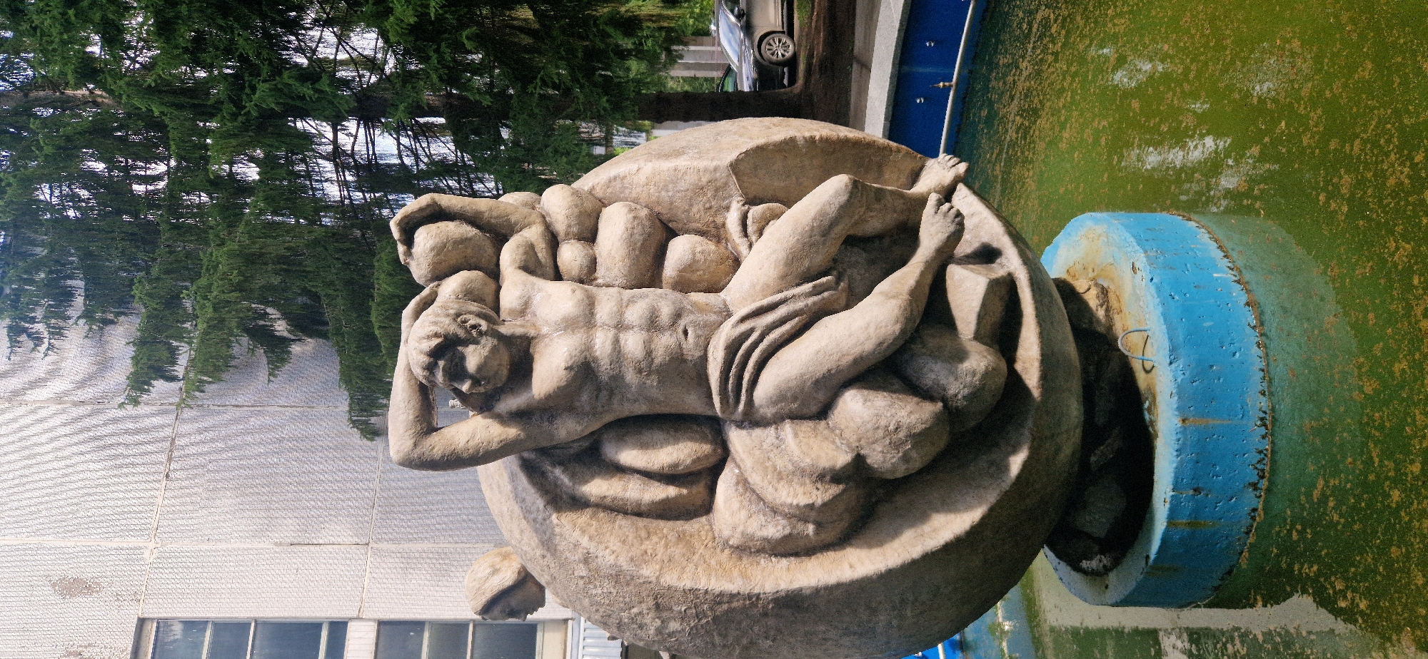 The image shows a stone sculpture of two figures in a fountain in the city of Krasnoobsk, Russia. The sculpture depicts a muscular male figure with his hands raised behind his head, supporting what appears to be a large stone sphere. To his left, a second, partially obscured figure, possibly female, is also leaning on the sphere. The sculpture is situated in a round fountain filled with murky green water. The base of the fountain is a blue, cylindrical structure. In the background, a modern building with a textured facade and a large window is visible, along with mature coniferous trees. A dark-colored car is parked on the right side of the frame, suggesting an outdoor urban setting. The lighting indicates it is daytime, with sunlight casting shadows on the sculpture.