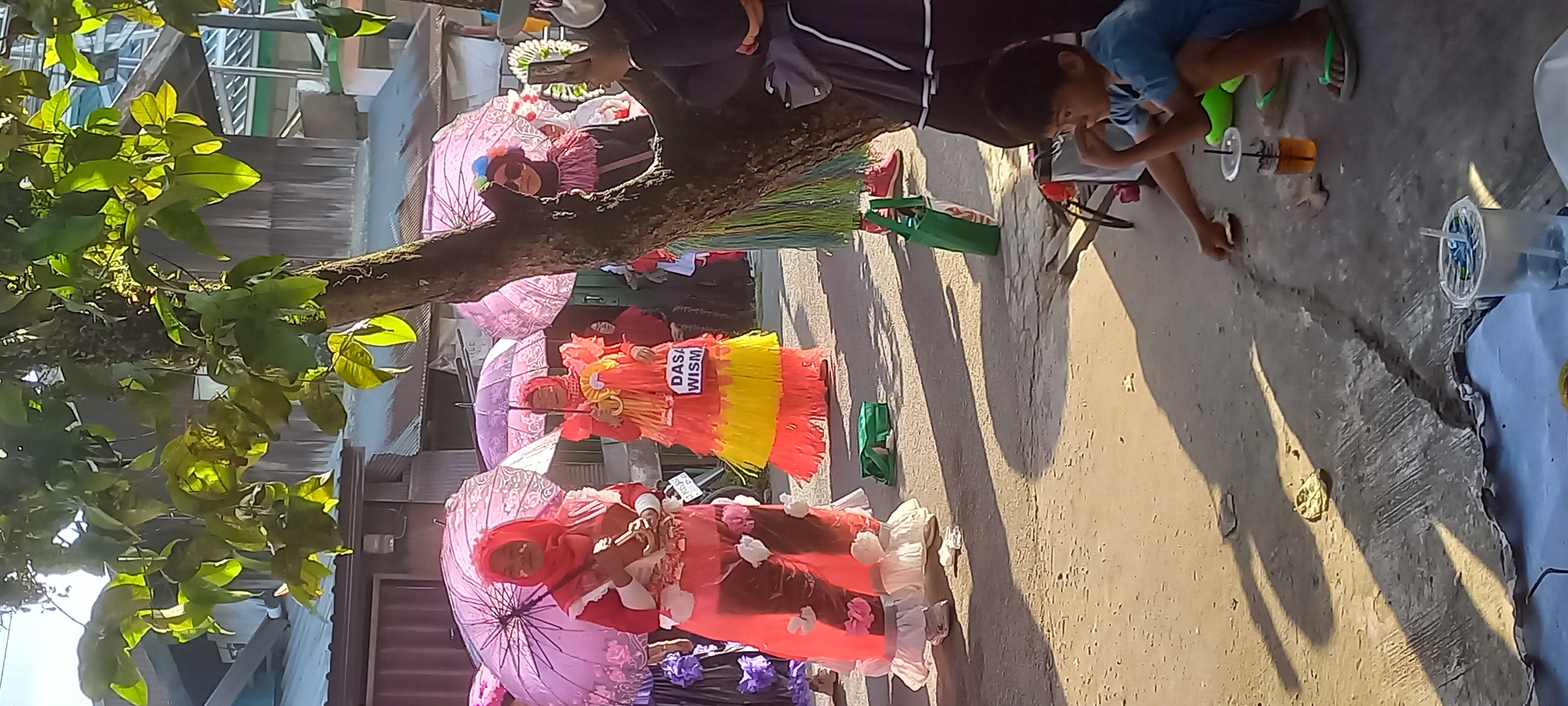 The image depicts an outdoor street scene, likely in Samarinda, Indonesia, given the context. In the foreground, a young boy in a blue t-shirt and green flip-flops is crouched on the ground, appearing to interact with something small. Two plastic cups with straws are visible nearby, one containing a dark liquid. The ground is paved with concrete and dirt, with shadows cast by trees and objects. In the mid-ground, three women are participating in what appears to be a parade or cultural event. They are wearing elaborate, brightly colored costumes, including flowing skirts and headscarves. Two of the women are holding pink umbrellas, and one has a sign attached to her costume that reads "DASA WISM". The overall atmosphere suggests a sunny day, with the sunlight creating distinct shadows. Buildings with corrugated metal roofs are visible in the background, characteristic of urban areas in Indonesia.