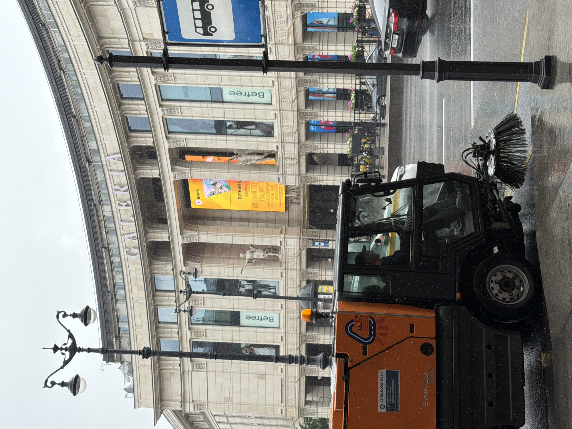 An orange and black street sweeping vehicle is positioned on the right side of the image, facing left and slightly towards the viewer. The vehicle has a driver inside, wearing a cap and a dark shirt. A large rotating brush is attached to the front underside of the vehicle, actively sweeping the wet pavement. The street is wet, indicating recent or ongoing rain.
Behind the street sweeper is a multi-story building with a light-colored facade. The building has a curved architectural design and features large windows, some of which display advertisements and signage. One prominent sign reads "GALERIA" in large letters across the top. Another large vertical sign is bright yellow and features Russian text. Smaller signs with various brand names are also visible.
In front of the building, a decorative black lamppost with multiple light fixtures stands prominently on the left side of the image. Another similar lamppost is positioned on the right side of the image, closer to the viewer, and a blue bus stop sign is attached to it.
Several vehicles, including a black sedan and a white car, are visible on the wet street in the background. People are sparsely scattered in the background, walking on the sidewalk in front of the building. The sky is overcast, contributing to the generally muted lighting of the scene. The overall impression is of a city street during a rainy day, with urban cleaning operations underway.