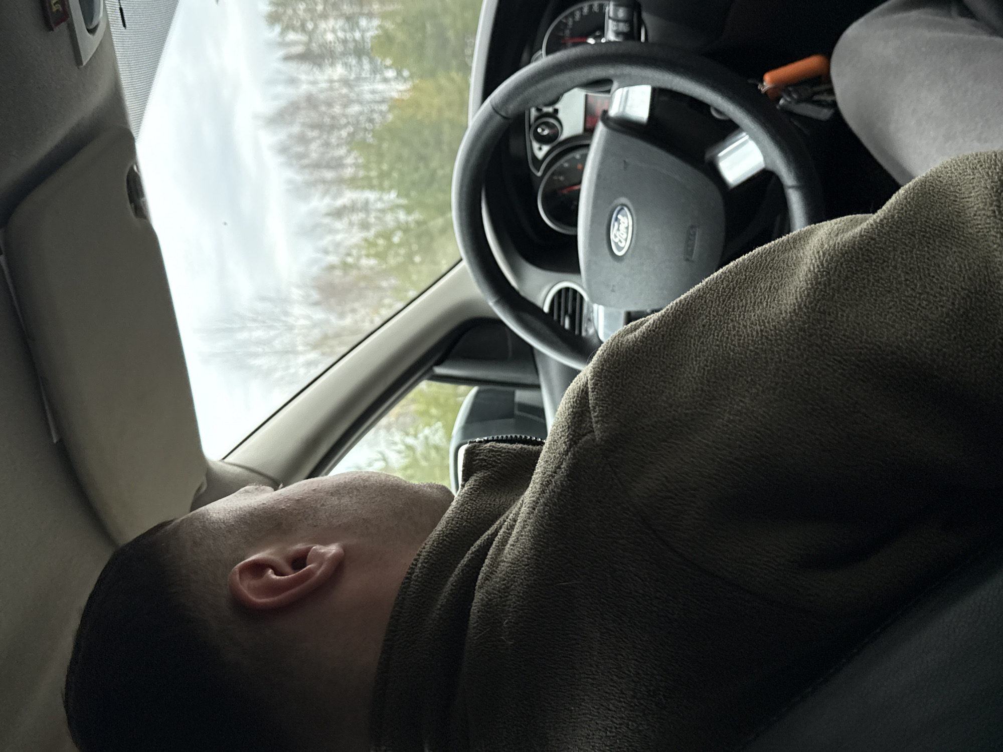 A man is slumped over the steering wheel of a Ford vehicle, presumably in Большая Уса, Russia, as the surrounding trees are visible through the windshield. The man has close-cropped hair and is wearing a dark, textured jacket. The car's interior is visible, including the dashboard, steering wheel with the Ford logo, and a partial view of the passenger seat. The sky is overcast.