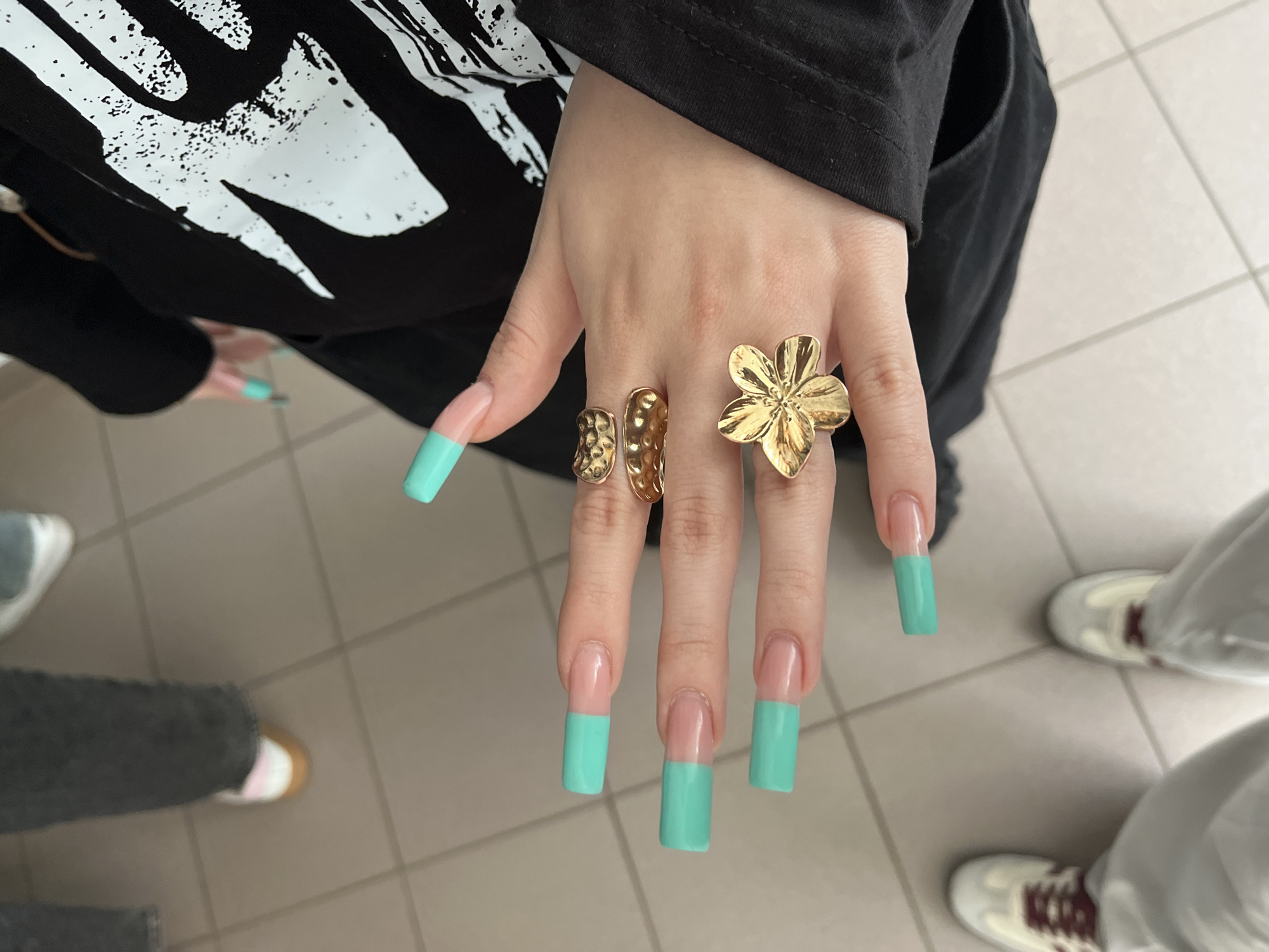 The image shows a close-up view of a person's hand, adorned with long, light pink acrylic nails with turquoise tips, and several gold rings. One ring features a floral design, another appears textured with small indentations, and a third is a simple, rounded band. The hand is resting on a black garment with a white graphic print. In the background, blurred feet wearing casual shoes and light-colored pants are visible, standing on a tiled floor. The scene appears to be indoors, possibly in a public space like a hallway or waiting area. There is no indication of the location being Sterlitamak, Russia.