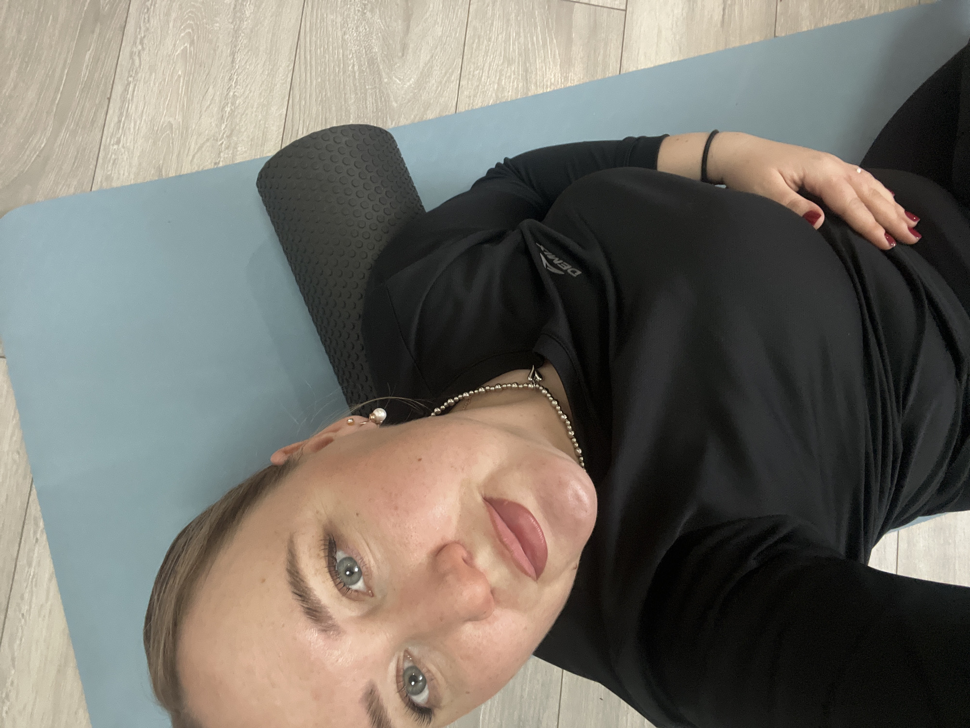 The image shows a young woman lying on her back on a light blue yoga mat, placed over a light-toned wooden floor. A black, textured foam roller is positioned horizontally beneath her upper back or shoulders. She is wearing a long-sleeved black athletic shirt and black leggings.
The woman has light skin, light-colored eyes, and light hair. Her lips are slightly parted and appear to have a light pink tint, and her fingernails are painted a dark red. She wears a silver-colored beaded necklace with a small dark charm, and small earrings, one of which looks like a pearl stud. Her left hand rests on her torso, and a black hair tie or band is visible on her left wrist.
She is looking directly at the camera with a neutral expression, suggesting she is taking a selfie. The activity appears to be related to fitness, stretching, or muscle recovery, utilizing the foam roller. The setting is indoors, with the wooden floor and yoga mat indicating a home or fitness space in Chaikovsky, Russia. On her left sleeve, the brand name "DEMIX" is visible in white lettering.