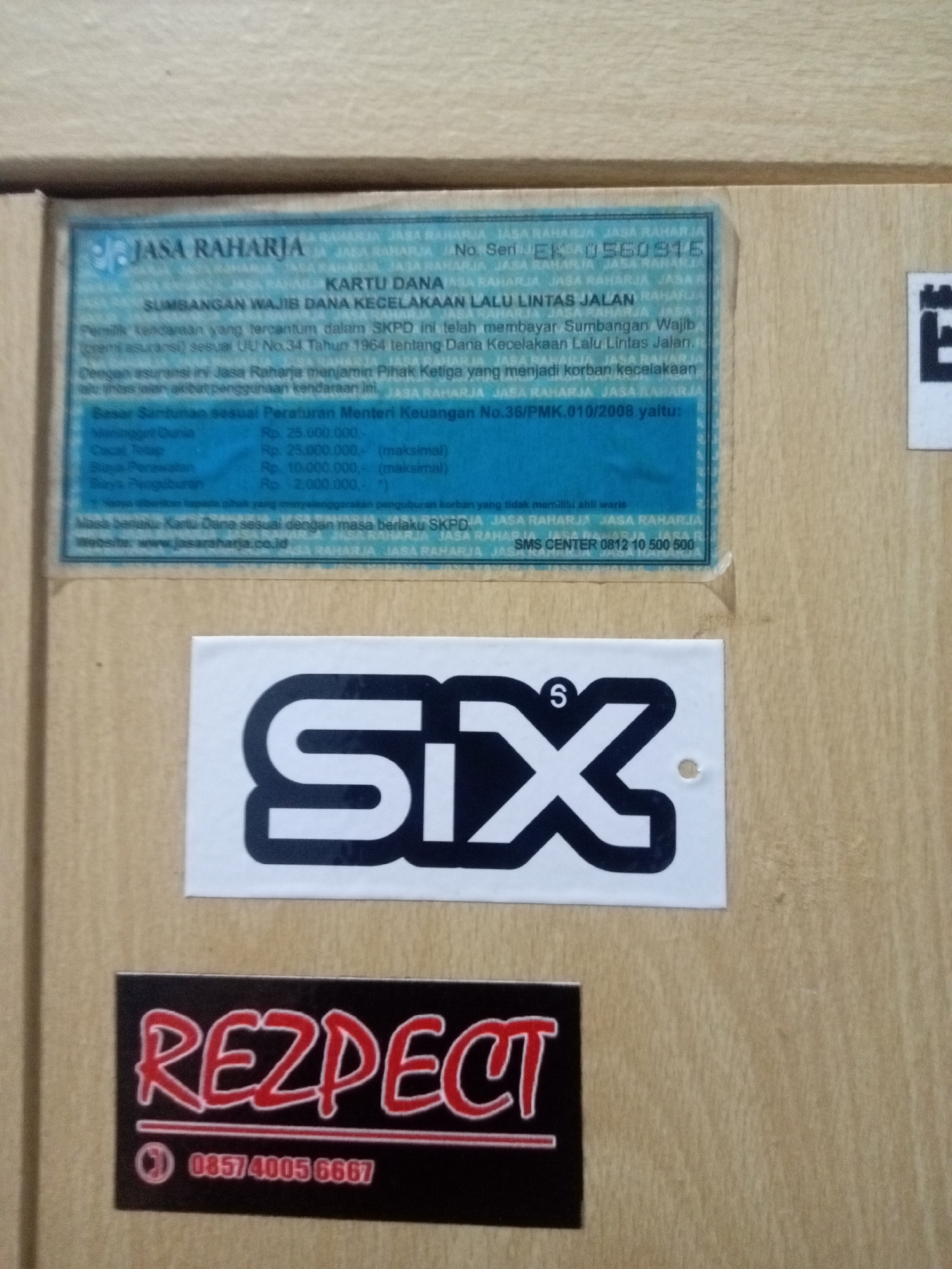 The image shows a close-up of a wooden surface with several stickers and a document adhered to it. The document is a blue and white card, likely an insurance or registration document, with text in Indonesian. The text includes "JASA RAHARJA," "KARTU DANA," and information about accident insurance. To the left of the document, there is a white sticker with a black stylized "SIX" logo. Below the document, there is a black sticker with red lettering that says "REZPECT" and a phone number, "0857 4005 6667." The lighting suggests an indoor environment, possibly a wall or a cabinet, with no discernible architectural features indicating a specific location in Batam, Indonesia. There are no people, actions, or interactions depicted in the image. The focus is solely on the objects themselves.