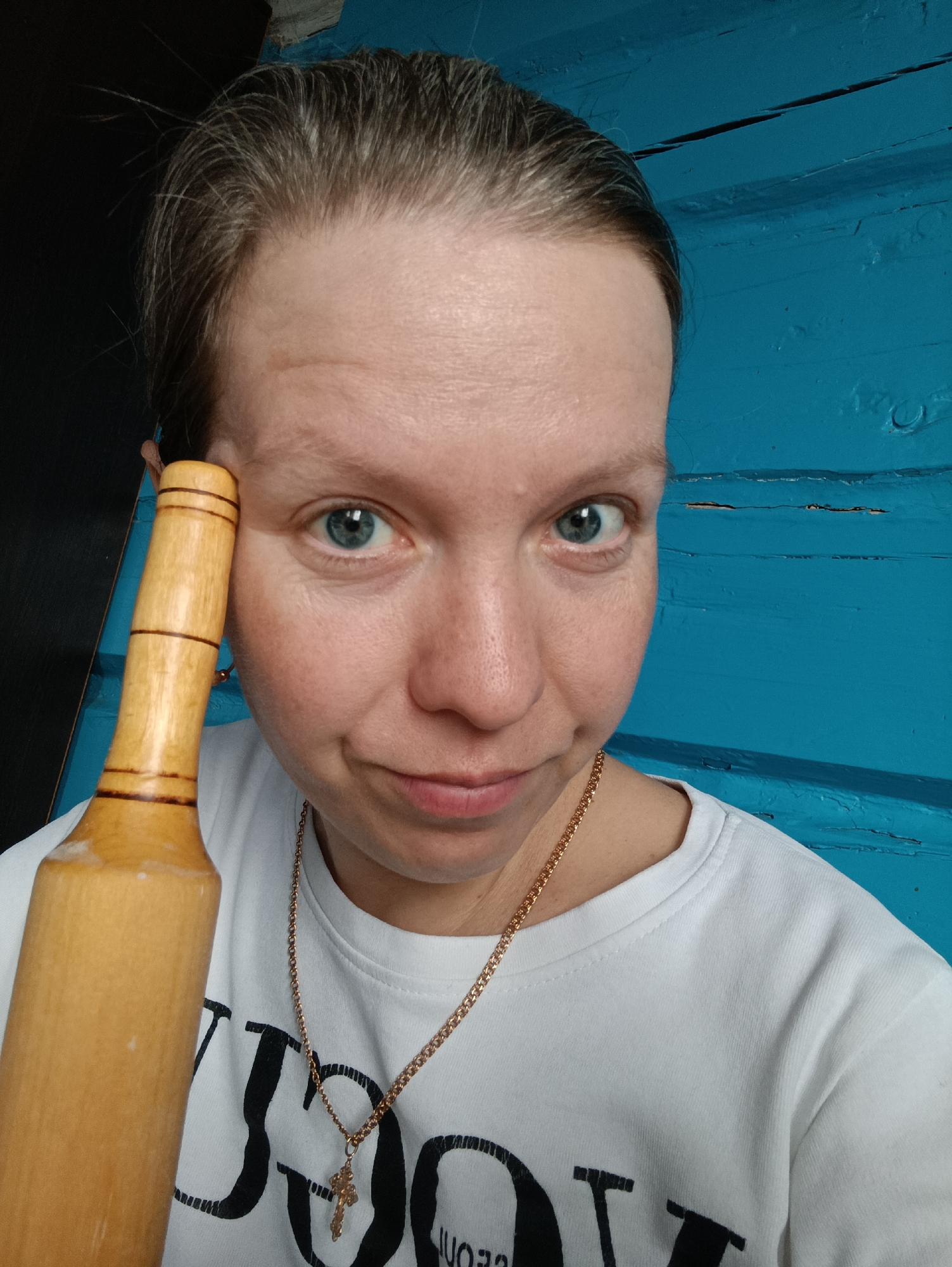 A woman is holding a wooden rolling pin next to her face. She is wearing a white t-shirt with black lettering and a gold chain with a cross pendant. The background is a blue wooden wall. The photo was taken in Пешнигорт, Russia.