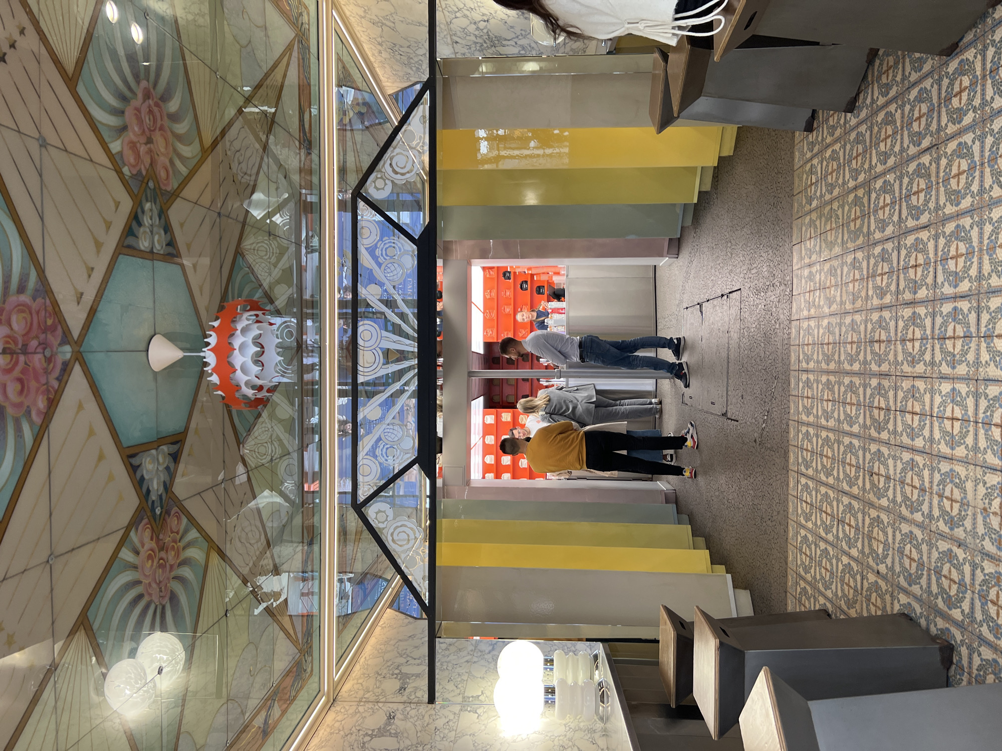 An elevated, rotated indoor shot captures a vibrant commercial space, likely a cafe or shop in Paris, France. The scene is dominated by an elaborate, possibly mirrored or glass, ceiling patterned with Art Deco-style geometric shapes and stylized floral motifs in shades of cream, light blue, pink, and green. A distinctive white and orange tiered light fixture hangs from the ceiling.
A prominent glass partition with black framing dissects the frame, reflecting parts of the interior. On the other side of this partition, several people are visible. Two men stand at a bright red counter or display at the back, one in a white shirt and jeans, the other in a mustard-yellow shirt and dark pants, seemingly engaged in an activity like ordering or waiting for service. Another person is partially visible behind them at the counter.
The wall on the right features a modern design, composed of horizontal rectangular panels in varying pastel and bright colors: white, beige, yellow, light green, light blue, and pink. The floor displays a classic pattern of blue and beige tiles with a repeating circular or star motif, transitioning to a darker, textured surface closer to the foreground. Minimalist, dark-colored seating elements and a table with glowing white lights are partially visible in the foreground. The overall impression is a blend of historic elegance (ceiling, floor) with modern, colorful accents. No discernible text is visible.