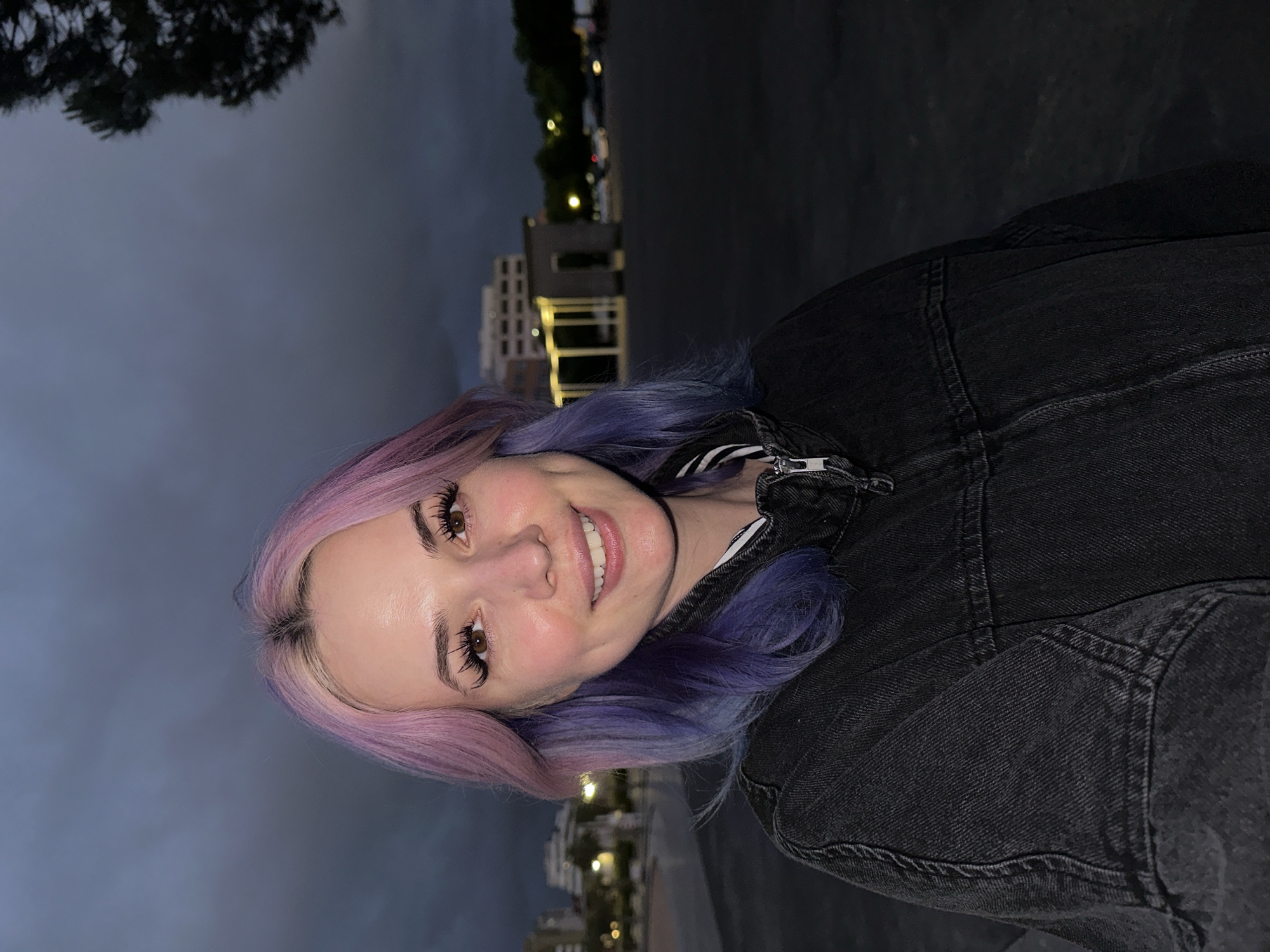 A cheerful young woman is captured in a close-up, waist-up portrait, smiling broadly at the camera. She has striking multi-toned hair, primarily shades of pink and purple, with some darker roots visible. Her eyes are open, revealing light brown irises framed by prominent, long false eyelashes and well-defined eyebrows. She wears a dark, possibly black or dark grey, denim jacket with visible seams and a metallic zipper detail on the front, over what appears to be a striped garment underneath. Her skin appears fair and smooth, with a subtle blush on her cheeks.
The setting is an urban environment at dusk or night in Gijón, Spain. The sky in the background is a deep, dark blue-grey, indicating low light conditions. Behind the woman, several multi-story buildings are visible, illuminated by scattered artificial lights that appear as soft glows. Trees, silhouetted against the dark sky, are also present in the upper left portion of the image. The overall atmosphere is dim but punctuated by the warm glow of city lights. No text is visible in the image.