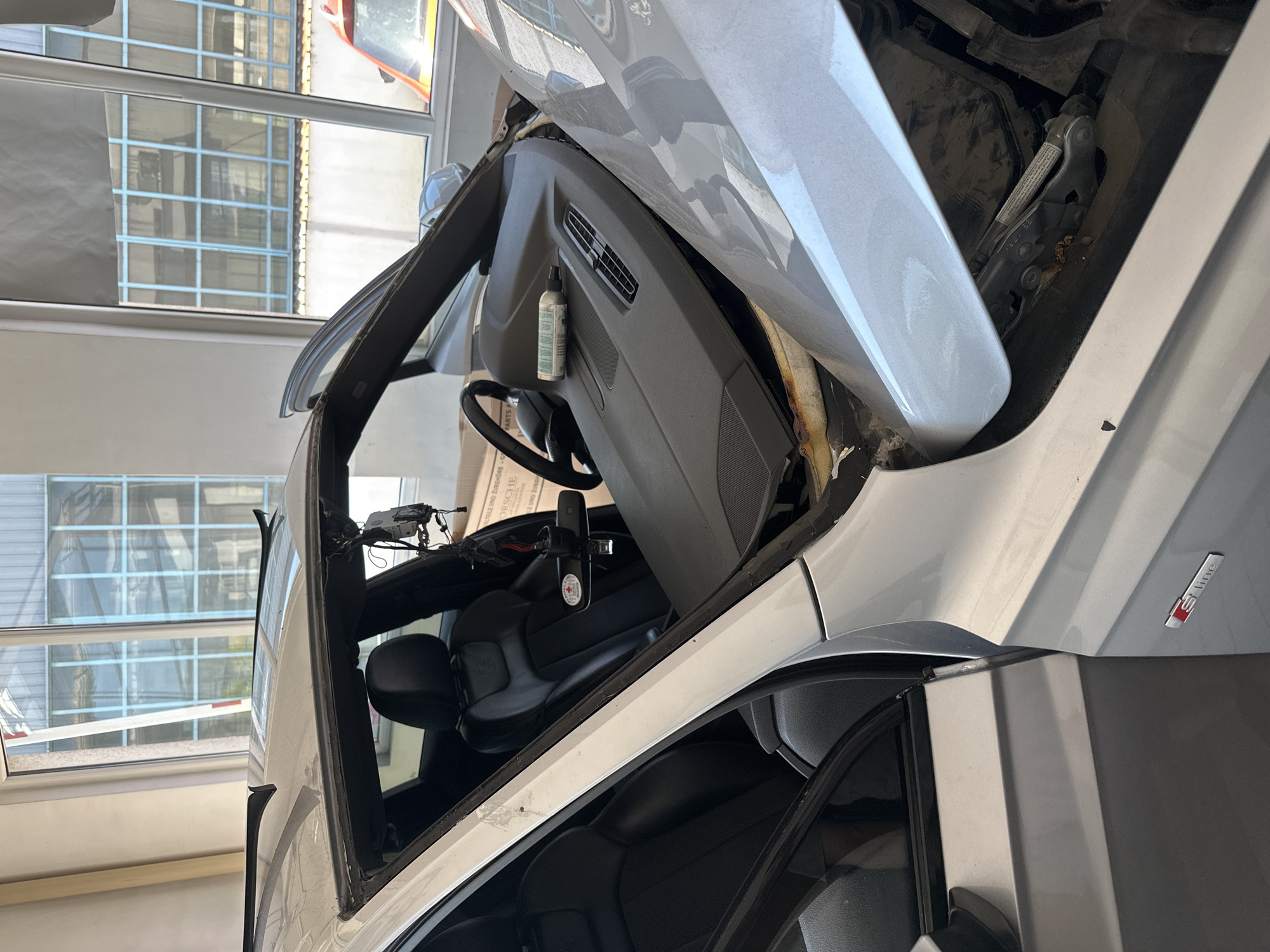 A silver Audi S-line is parked indoors, likely in an auto repair shop, with its hood and driver's side door open. The windshield has been removed, revealing the interior of the car, including the dashboard, steering wheel, and black leather seats. A can of sealant is placed on the dashboard. The car appears to be undergoing maintenance or repair. The background shows windows with a view of a building with blue grid-like structures.
