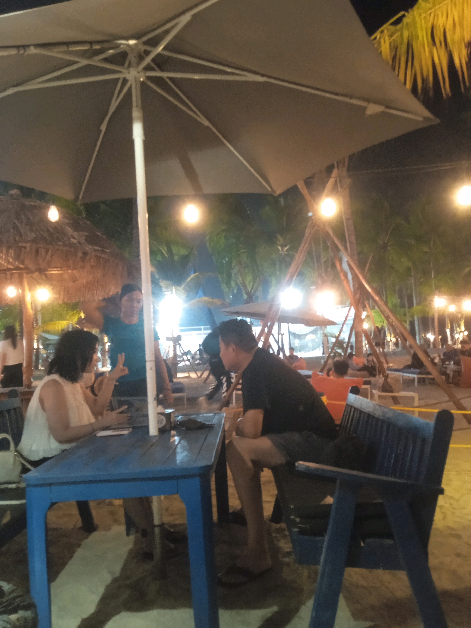 The image depicts an outdoor dining scene at night, likely in Kosambi Barat, Indonesia, given the presence of palm trees and a generally tropical ambiance. The foreground is dominated by a blue table and chairs, with two individuals seated at the table. A woman in a white top is on the left, gesturing with her left hand. A man in a black t-shirt is on the right, facing her. Behind them, another person in a teal shirt stands near the table.
The setting appears to be a beachside or resort area. Large umbrellas provide shade, and string lights illuminate the area, creating a warm, inviting atmosphere. In the background, more seating areas are visible, with other patrons enjoying the evening. The sand underfoot and the lush vegetation suggest a coastal location. The lighting is dim, with several artificial light sources creating reflections on the table.