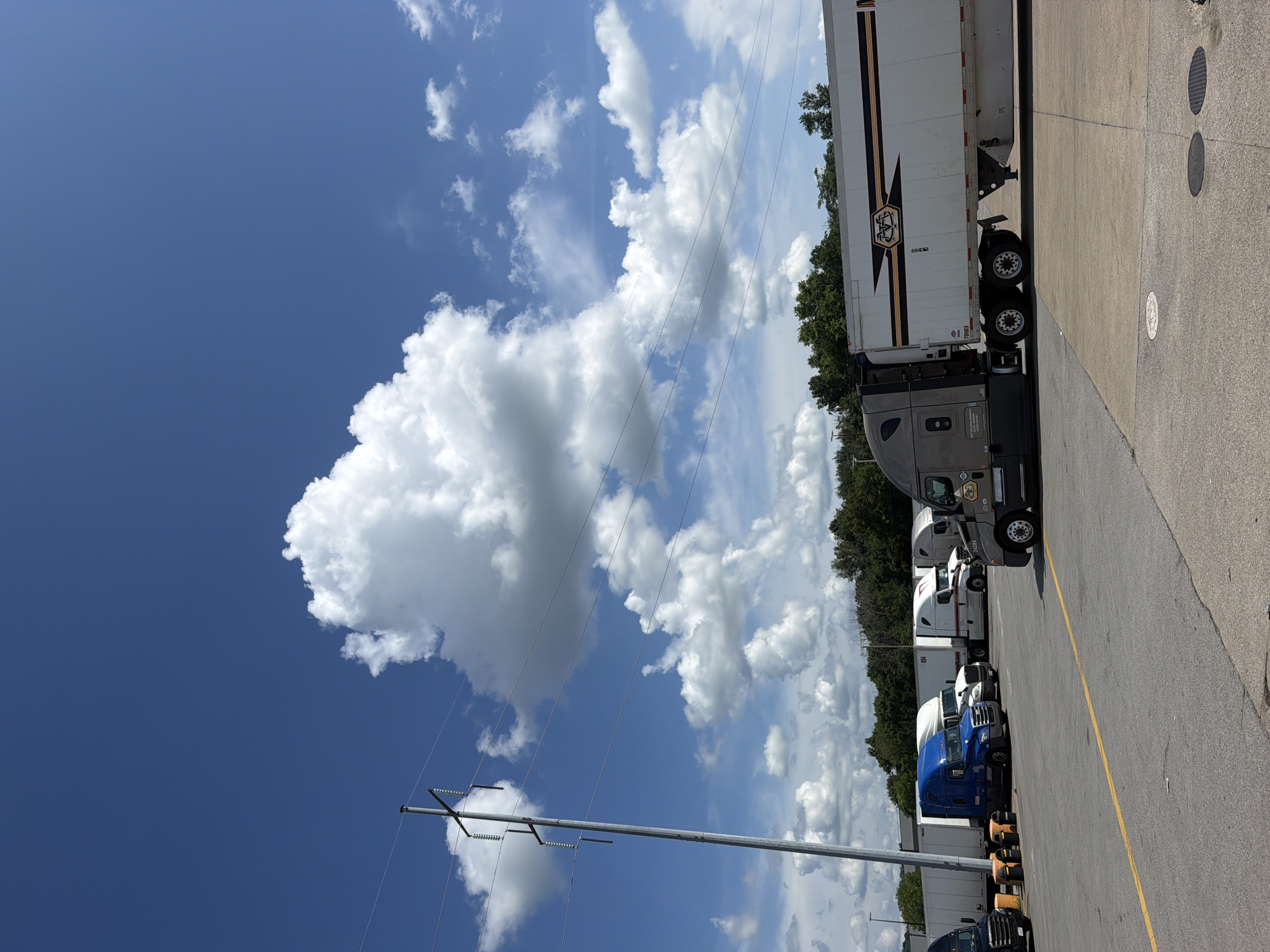A sunny daytime, low-angle exterior shot captures a parking lot filled with commercial trucks and trailers under a bright blue sky dotted with large, white cumulus clouds. A prominent utility pole with multiple wires stands to the left of the frame. Several semi-trucks, some blue, some white, and a dark grey one in the foreground, are parked in rows. The grey truck, a Freightliner model, is equipped with a white trailer featuring a distinctive black and orange logo and stripe design. The parking lot surface is asphalt with yellow line markings. In the background, a line of green trees forms a treeline. The scene is consistent with a truck stop or a depot.