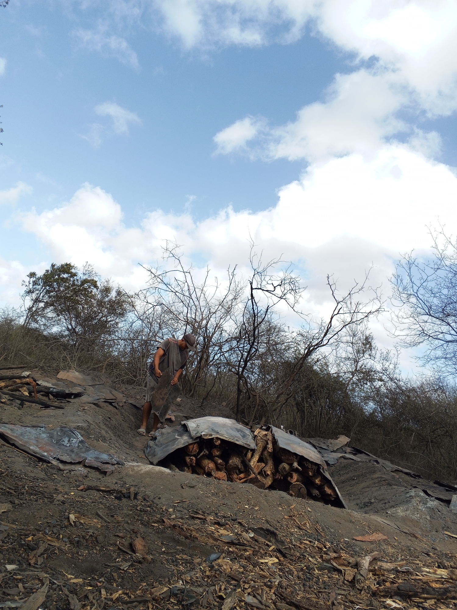 The image shows a scene in Altagracia, Venezuela, depicting the aftermath of a fire. In the foreground, there's scorched earth and debris, along with charred wood. A man stands amidst the wreckage, dressed in a t-shirt, shorts, and a baseball cap, appearing to be sifting through the rubble. There are pieces of corrugated metal, some covering stacks of firewood, scattered around the area. The background consists of bare trees and shrubbery, silhouetted against a cloudy sky.