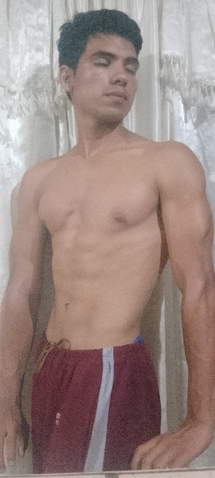A young adult male is depicted in a medium-close-up shot, standing against a light-colored, vertically textured fabric background. The subject is bare-chested, displaying a muscular physique, and wears maroon shorts with a single light-colored vertical stripe on the viewer's left leg. His dark, short, curly hair is visible. His eyes are closed or heavily lidded, and his head is slightly turned to the viewer's right. The subject's left hand is visible at the bottom right of the frame, resting on a flat, horizontal surface. The scene is located in Juan Griego, Venezuela.