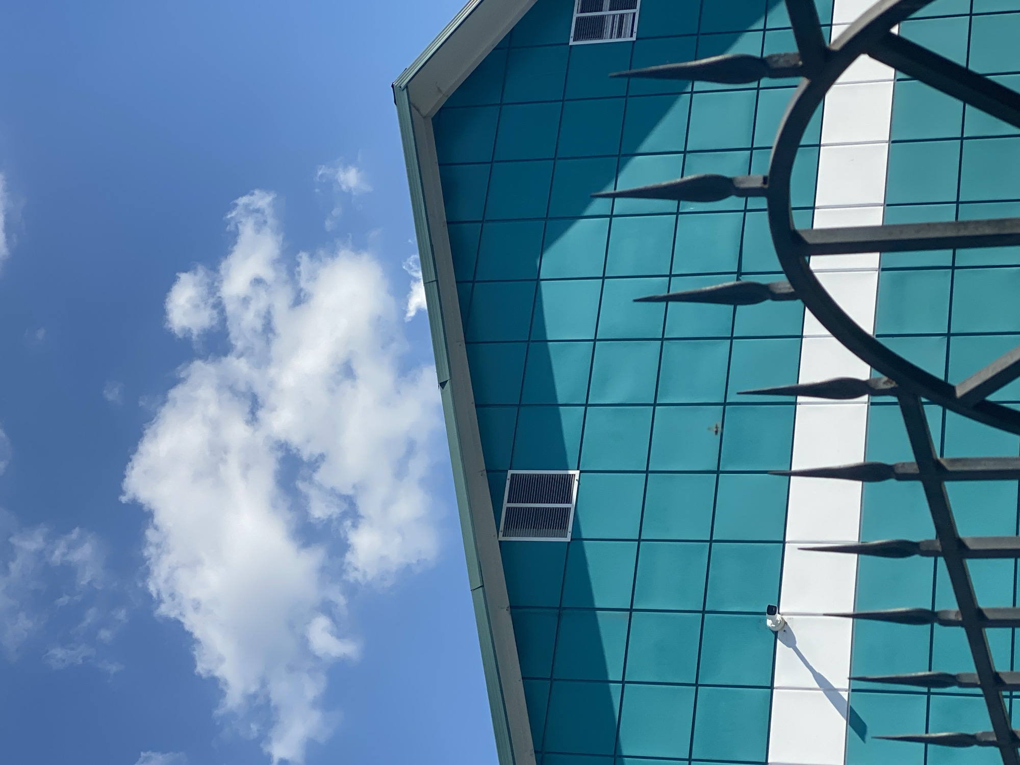 The image shows the facade of a modern building with teal-colored panels and a white horizontal stripe. A black metal fence with pointed finials is in the foreground, partially obscuring the view of the building. The building has two small, rectangular windows with louvers. A security camera is visible mounted on the wall. The sky is blue with scattered white clouds. The scene is outdoors on a sunny day. There are no people or animals visible in the image. The location is not specified as Novokuznetsk, Russia.