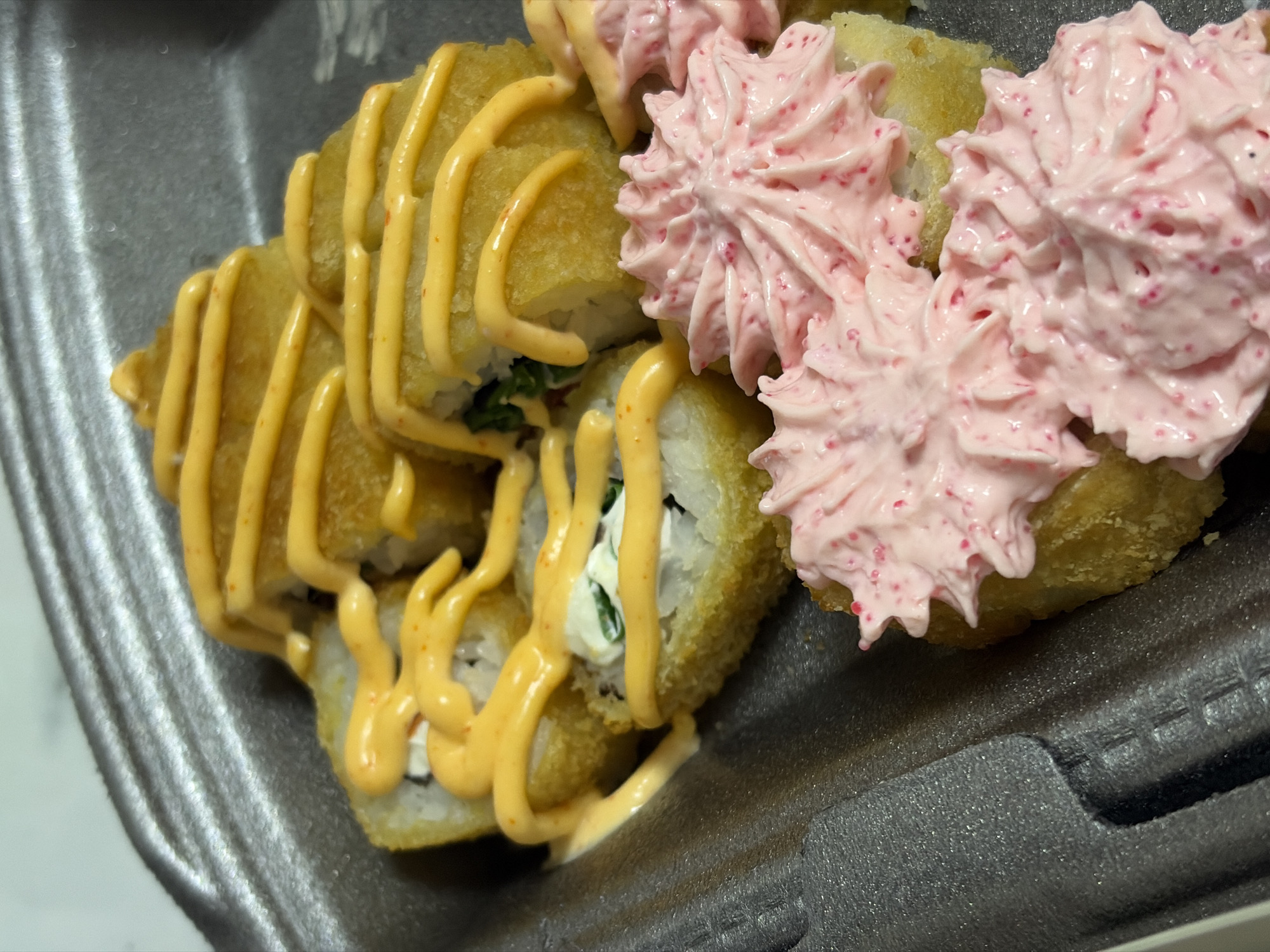 The image shows a take-out container of sushi. One set of sushi rolls is deep-fried and topped with a drizzled orange sauce. The other set is topped with piped pink cream with red specks. The container is gray and appears to be made of styrofoam. Location is unknown.