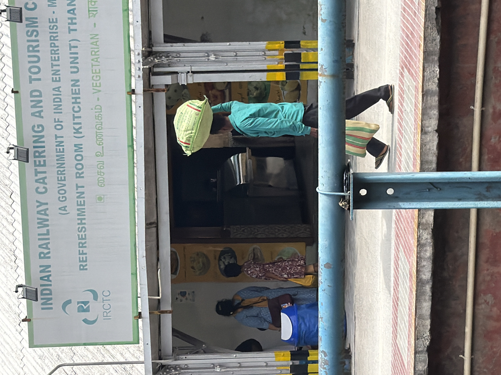 The image captures a scene at what appears to be a train station in Thanjavur, India. A prominent sign displays "INDIAN RAILWAY CATERING AND TOURISM C" and "IRCTC (A GOVERNMENT OF INDIA ENTERPRISE - M REFRESHMENT ROOM (KITCHEN UNIT) THAN VEGETARIAN - शाक".
In the foreground, a person in a teal-colored shirt and dark pants is lying down, possibly resting or sleeping, with a light green bag acting as a pillow. Their legs, clad in dark pants and flip-flops, are visible, as is a colorful striped bag placed near their feet.
Further back, through an opening, two other individuals are partially visible. One person appears to be lying down, while another is seated or in a similar resting posture. The background suggests an indoor area, perhaps part of the refreshment room, with posters or artwork on the walls. The overall lighting suggests it is daytime. The presence of the IRCTC sign and the visual of people resting strongly indicate a railway station environment. The scene conveys a sense of everyday life and rest in a public transit hub.