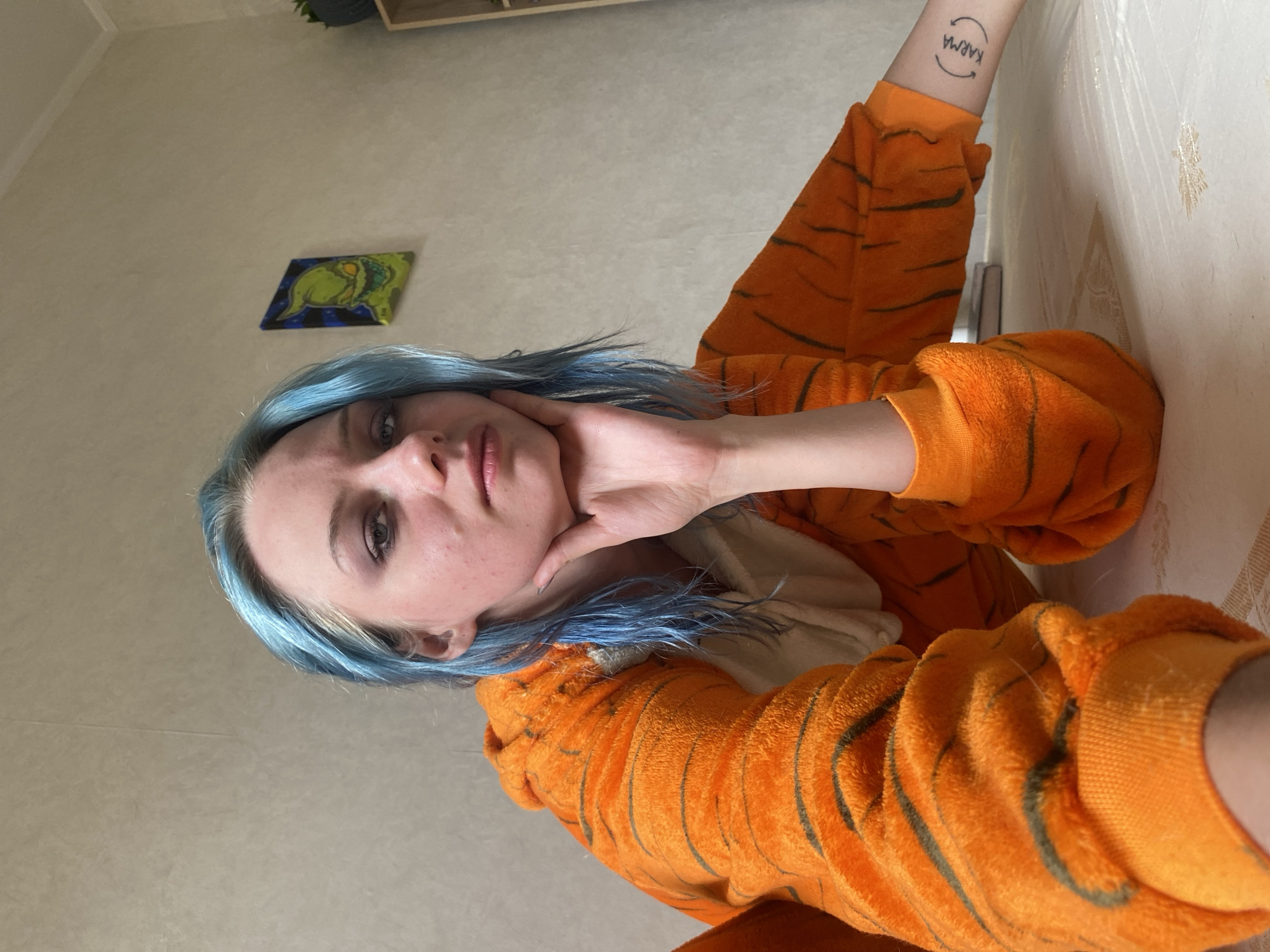 A young person with blue and blonde hair is captured in a close-up, waist-up shot, leaning forward and resting their chin on their hand. They are wearing an orange onesie with black tiger stripes. The background features a textured wall with a small framed picture of a green character on it. The scene appears to be indoors, possibly a home environment, with no discernible elements placing it specifically within Bishkek City, Kyrgyzstan. The lighting is neutral, suggesting daytime.