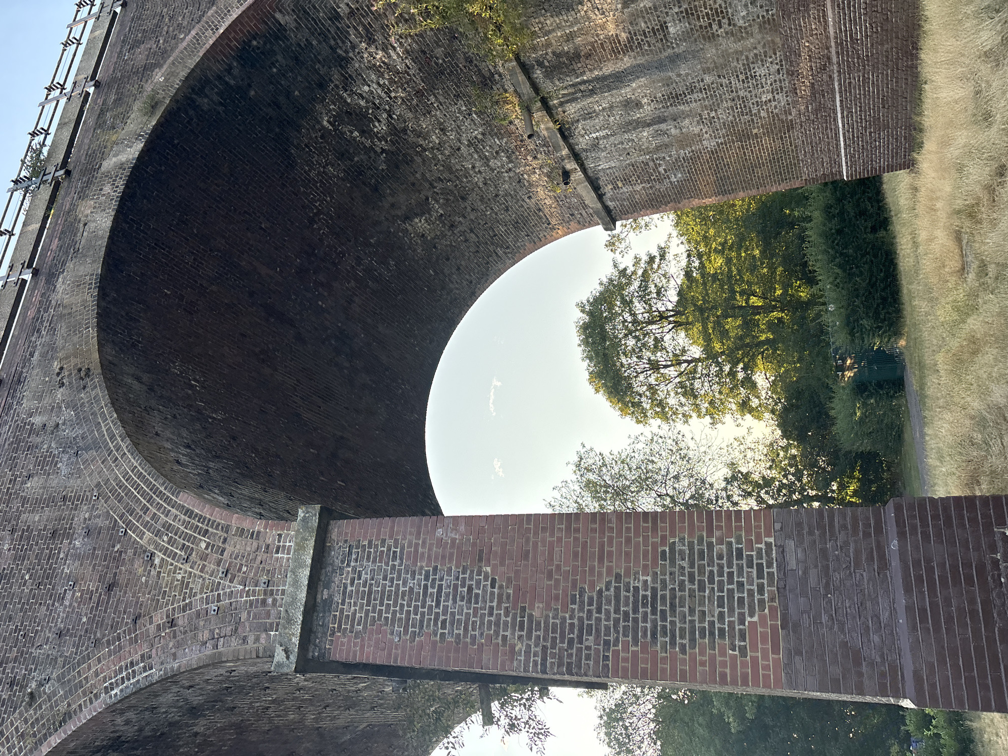 The image shows a low-angle view of a brick railway viaduct, possibly the Chelmsford Railway Viaduct, in Chelmsford, United Kingdom. The viaduct has multiple arches, with the perspective focused on the underside of two adjacent arches. The bricks are weathered, with some areas showing signs of repair or different brick types.
Supporting the viaduct on the left is a substantial brick pillar, showing a diagonal pattern of darker bricks against the reddish-brown brickwork. The pillar appears to be in good structural condition. The arches rise up and disappear out of the frame, with the sky visible through the gaps.
Beyond the viaduct, a lush green landscape is visible, with trees and bushes. The ground in the foreground is covered in dry, golden grass. Sunlight casts long shadows, indicating it is either early morning or late afternoon. The sky is a clear, pale blue with a few wispy clouds. There are no people or vehicles clearly visible in the immediate scene, although there may be distant figures in the background.
The overall impression is of an old, functional piece of industrial architecture integrated into a natural setting.