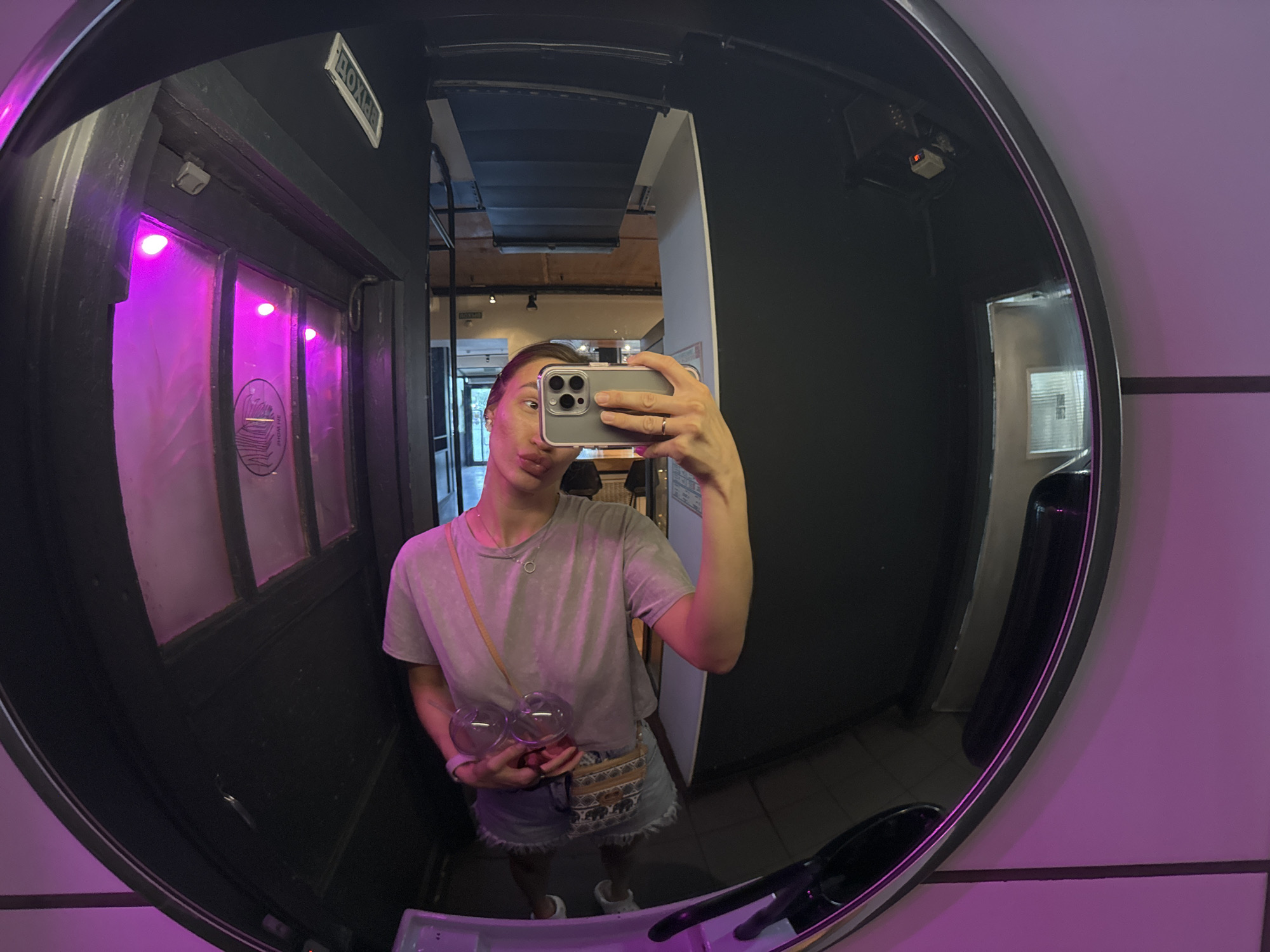 A person is taking a selfie in a circular mirror. The person is wearing a gray t-shirt, denim shorts, and white sneakers, and is holding two wine glasses. The background includes a dimly lit interior with black walls and a door with a sign in Russian, indicating "ВЫХОД" (Exit). The location appears to be a bar or restaurant in Nizhny Novgorod, Russia. The overall lighting is moody, with pink hues adding to the atmosphere.