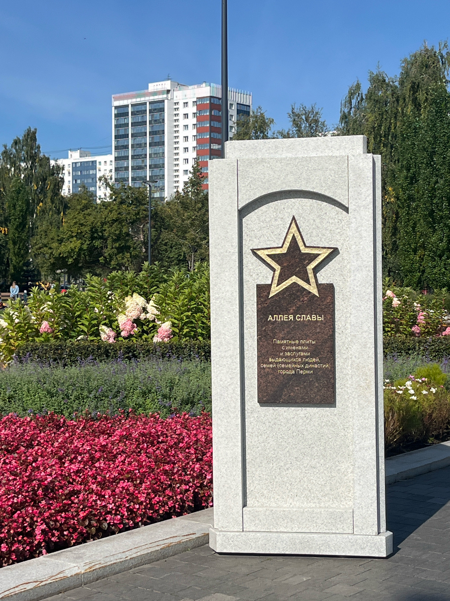 A monument with a star and an inscription stands in a park. The inscription reads "АЛЛЕЯ СЛАВЫ" (Alleya Slavy, meaning "Avenue of Glory"). Below this, it states "Памятные плиты с именами и заслугами выдающихся людей, семей (семейных династий) города Перми" (Memorial plaques with the names and merits of outstanding people, families (family dynasties) of the city of Perm). The monument is made of light granite and features a bronze plaque with a star emblem. Behind the monument, there are lush green trees and flowering bushes in shades of pink and white. In the background, there are modern residential buildings. A paved walkway runs in front of the monument. The sky is clear and blue, indicating a sunny day. There are a few people visible in the distance, appearing to be walking or sitting in the park. The overall scene depicts a public space dedicated to honoring notable individuals and families from the city of Perm.