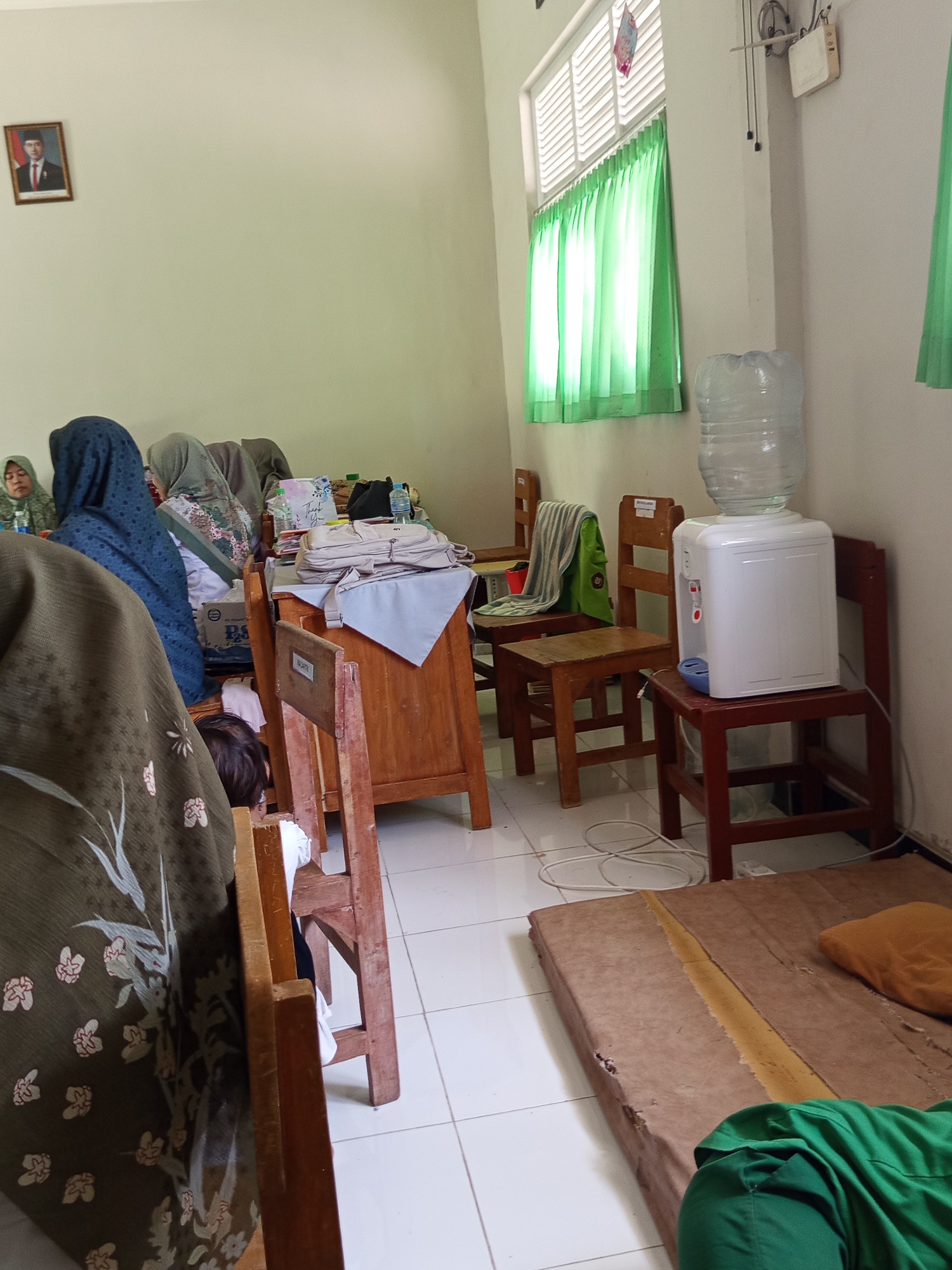 The image depicts an indoor scene, possibly a classroom or communal space, in Lumutan, Indonesia. The room is sparsely furnished with wooden chairs and tables. A water dispenser is present, indicating a likely public or shared space. Several individuals, predominantly women wearing headscarves, are visible in the background, engaged in what appears to be a gathering or meeting. One individual is seated at a desk, while others are positioned around tables. A framed portrait of a man in official attire hangs on the wall, suggesting a connection to a government or educational institution. The overall atmosphere appears casual and informal, with evidence of everyday activities such as the presence of bags and personal items. The flooring is tiled, and the walls are painted a light, neutral color. Green curtains are visible, suggesting a window or entrance. A mattress lies on the floor in the foreground, along with some clothing items, hinting at a temporary or makeshift sleeping arrangement. The lighting suggests it is daytime, with light filtering in from a window.