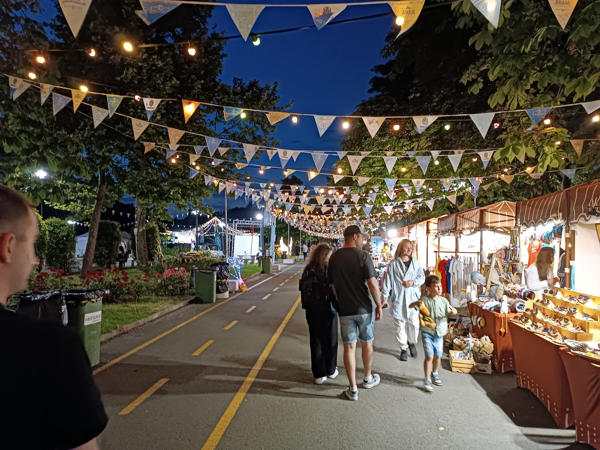 This detailed description outlines an outdoor evening scene, likely a market or festival, within the Sea Garden (Morska Gradina) in Burgas, Bulgaria. The identification of Burgas is supported by numerous triangular pennants strung overhead, which prominently display the city's name "BURGAS" along with the coat of arms for "ОБЩИНА БУРГАС" (Municipality of Burgas) and the year "2021" on some.
The image is captured from a slightly elevated perspective, looking down a wide, paved pathway during twilight or night, evidenced by the deep blue sky. Overhead, multiple parallel lines of triangular pennants and spherical lights are suspended between the leafy canopies of mature trees. The pennants are predominantly white, light blue, and light yellow/beige, bearing civic branding. The spherical lights emit a warm, yellowish-orange glow, illuminating the scene.
The paved pathway, dark grey in color, features two parallel sets of interrupted yellow lines, suggesting designated lanes for pedestrian or bicycle traffic.
To the left of the path, a landscaped area contains green grass, dense green bushes, and beds of flowering red roses. Two green waste bins are visible along this strip; one has a black liner protruding and displays "EVENTSURCE" on its side. Further in the background on the left, white tent structures, scaffolding, and event lighting are partially visible.
To the right of the path, a continuous line of market stalls extends into the background. Each stall features a brown and white striped canopy or awning. Merchandise is displayed on counters and tiered wooden racks. Products visible include numerous pairs of sunglasses or similar items, various garments hanging on racks, and plush toys, including ducks and whales. At least two individuals, presumably vendors, are partially visible behind the stalls, one wearing a white long-sleeved top.
Several people are present on the pathway. In the immediate foreground, the back of a person's head and left shoulder, wearing a black t-shirt, is partially cropped on the left. Further down the path, a woman with curly dark hair, dressed in dark clothing and a backpack, walks away from the viewer, accompanied by a man in a dark t-shirt, denim shorts, and a dark baseball cap. Approaching the viewer are a woman with blonde hair, wearing a light-colored long jacket over white pants, and a young boy next to her, dressed in a light green t-shirt and denim shorts, holding a small brown stuffed animal in his right hand.