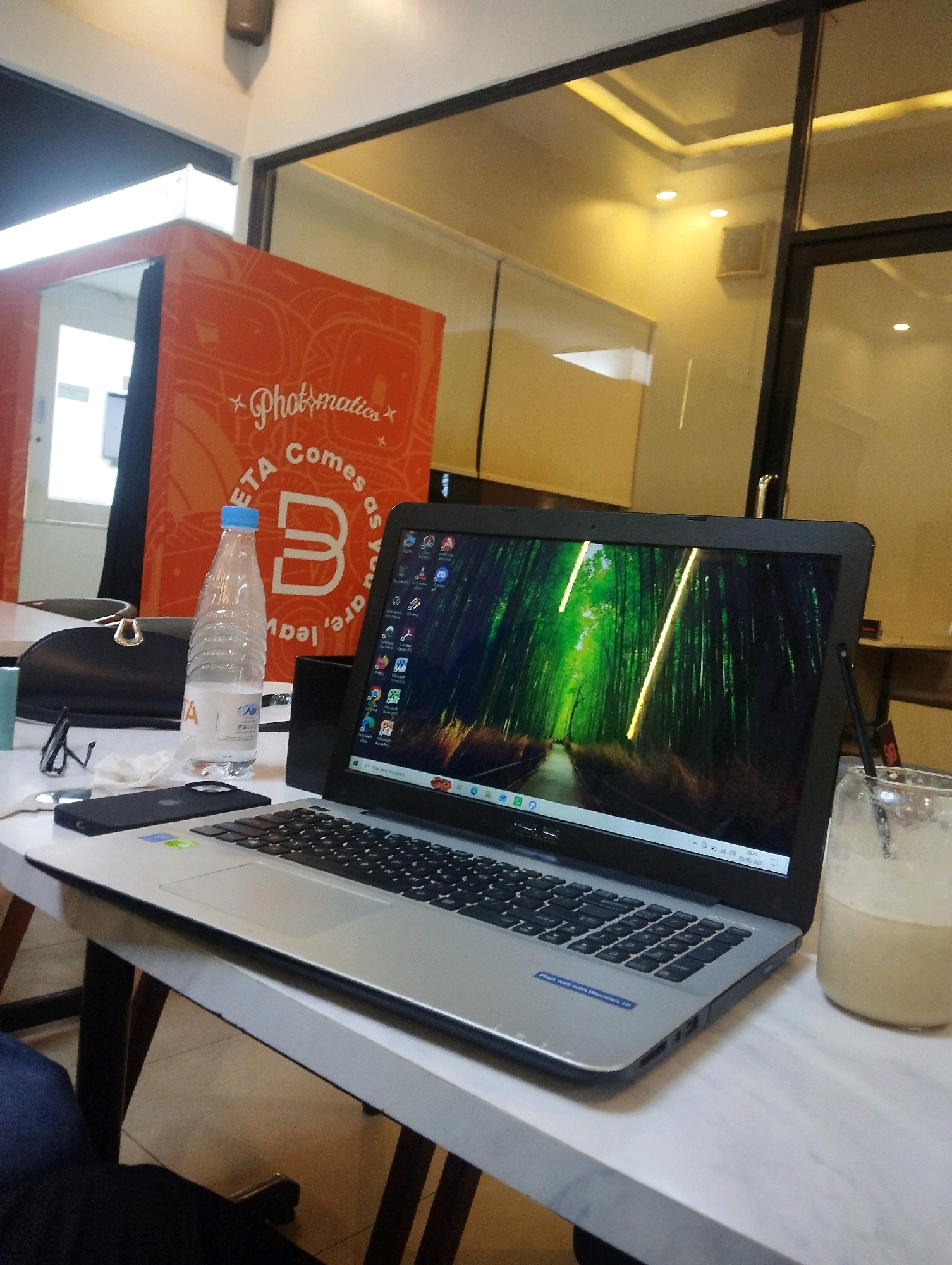 In a dimly lit indoor setting, likely a co-working space or cafe in Pekanbaru, Indonesia, a laptop is open on a white table. The laptop screen displays a vibrant wallpaper of a bamboo forest with yellow light filtering through. On the table, to the left of the laptop, sits a clear plastic bottle of water with a blue cap, a black purse, and a pair of black-rimmed glasses. To the right of the laptop is a glass containing a light-colored beverage with a straw. In the background, a large orange structure with white lettering that reads "Photomatics" is visible. The environment appears to be near a glass partition, suggesting a modern, possibly open-plan office or commercial space. The lighting is warm, casting a yellow hue on the ceiling and walls. No individuals are clearly depicted in the image, though the presence of personal items suggests a user was recently present or is nearby.