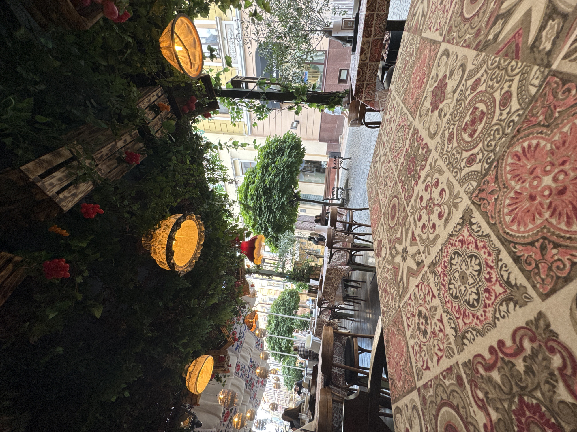 The image captures an outdoor seating area of a cafe or restaurant, likely located in Chernivtsi, Ukraine, given the architectural style of the buildings visible in the background. The foreground features a table covered with a patterned tablecloth, displaying a mosaic of floral and geometric designs in shades of beige, pink, and brown. Beyond this table, rows of tables and chairs are set up on a cobblestone-like surface.
The seating area is covered by a canopy formed by greenery and wooden structures, with several decorative lamps hanging from it, emitting a warm, orange light. Some of these lamps are shaped like ornate lanterns with lace trim, while others are simpler. Artificial grape clusters are also suspended from the canopy, adding to the rustic, decorative theme.
In the background, a tree with a rounded canopy stands prominently, and beyond that, buildings with yellow and beige facades are visible, suggesting an urban setting. Several people are seated at tables in the middle ground, appearing to be patrons of the establishment. Some are engaged in conversation, while others are looking around or have their backs to the camera. The overall atmosphere appears relaxed and inviting, typical of an outdoor dining space.