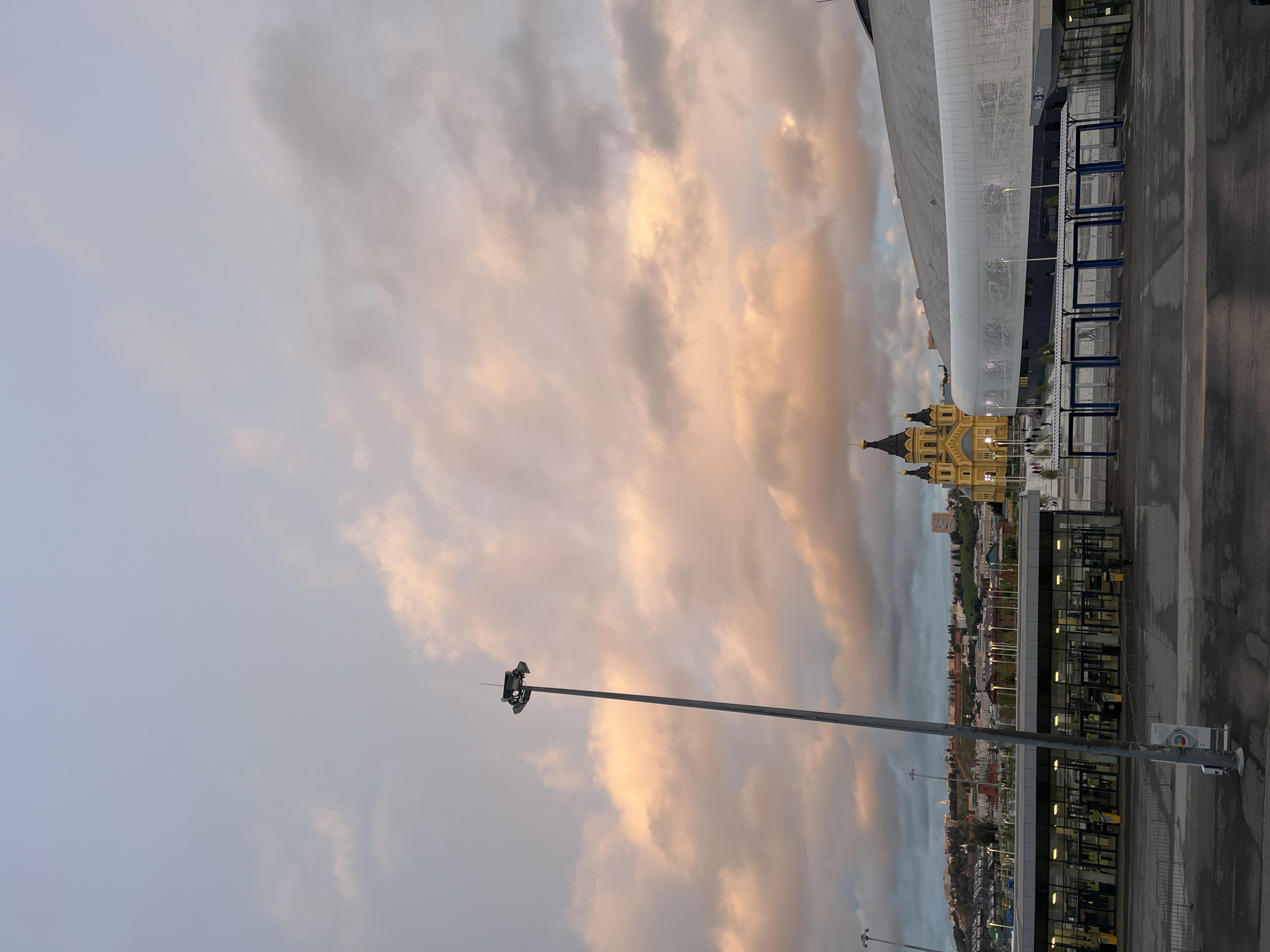 The image captures an exterior view of the Nizhny Novgorod Stadium in Nizhny Novgorod, Russia, during what appears to be sunset or dawn. In the foreground, a modern stadium structure with a white, curved facade is visible. To the left of the stadium, a bright yellow cathedral with multiple domes and spires stands out against the sky.
In front of the stadium, there is a walkway with several entrances, marked by turnstiles and covered waiting areas with blue metal frames. A tall lamppost with multiple lights is positioned prominently in the foreground, slightly to the left of center. In the middle ground, beyond the stadium and cathedral, a cityscape with buildings and hills is discernible. The sky is filled with dramatic clouds, some tinged with pink and orange hues from the setting or rising sun, contrasting with cooler gray tones. The ground in the foreground is wet, suggesting recent rain. The overall scene is dominated by the architecture of the stadium and cathedral, set against a dynamic sky.