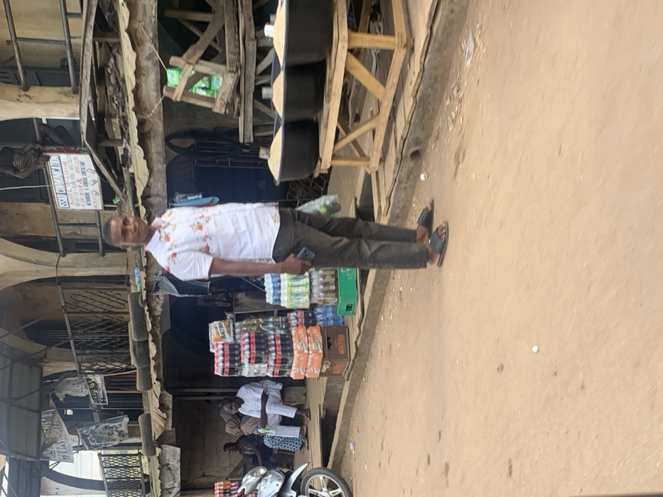 The image captures a bustling street market scene in Egbedi, Nigeria, under bright daylight. In the foreground, a dark-skinned male, approximately 25-35 years old, stands facing slightly left of the camera. He wears a white short-sleeved collared shirt with a red floral pattern, dark brown trousers, and black slide sandals. He holds a dark-colored smartphone in his left hand and a clear plastic bag containing green contents in his right hand.
To the left of the central figure, a silver motorcycle's front portion is visible. Behind it, a commercial display features tall stacks of bottled beverages, including red-labeled soft drinks in yellow plastic crates and blue-labeled water bottles in green plastic crates. Further left and behind these products, three individuals are seated on a raised concrete ledge: one male in a white traditional garment holds a small object, flanked by another male to his left, and a partially obscured figure further left, possibly wearing a head covering.
To the right of the central figure, a wooden market stall displays three large black tubs, each filled with a light-colored, granular substance. Behind these stalls, the street is lined with weathered, multi-story buildings featuring arched windows and balconies, indicative of commercial establishments. A white sign with "OSSAD COMMUNICATION" is visible on one of these buildings. Makeshift roof structures, constructed from wood and corrugated metal, are present, with several stacked tires observed on one lower roof. The ground surface is unpaved, composed of reddish-brown dirt or sand, with a concrete drainage gutter running parallel to the storefronts. A small white object lies on the ground near the gutter. No direct interactions or specific actions between the individuals are discernible.