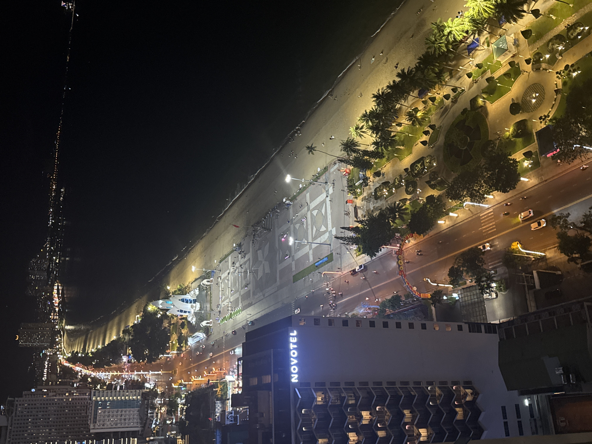 This high-angle, nighttime aerial view captures a lively coastal city scene. In the foreground, a prominent multi-story building, identified by the illuminated "NOVOTEL" sign on its side, stands out with a modern architectural design featuring distinctive patterned balconies or windows.
Adjacent to the building and running parallel to the coastline is a brightly lit, multi-lane road. Vehicles, including cars and motorbikes, can be seen in motion, with some leaving light trails, indicating the time of day. Beyond the road lies a wide, elaborately landscaped park or esplanade, lush with numerous palm trees and other greenery. The park is extensively illuminated by streetlights and decorative ground lighting, revealing pathways, garden beds, and scattered small figures of people strolling and congregating.
Further along, bordering the park, is a sandy beach. The gentle surf line is visible as waves meet the shore. Numerous individuals are seen walking along the water's edge and on the sand, enjoying the evening. The dark expanse of the ocean stretches into the background, with a distant line of lights visible on the horizon, possibly from other coastal structures or vessels.
The overall scene depicts a bustling urban waterfront at night, characterized by vibrant artificial lighting and considerable human activity, suggesting a popular leisure or resort area.