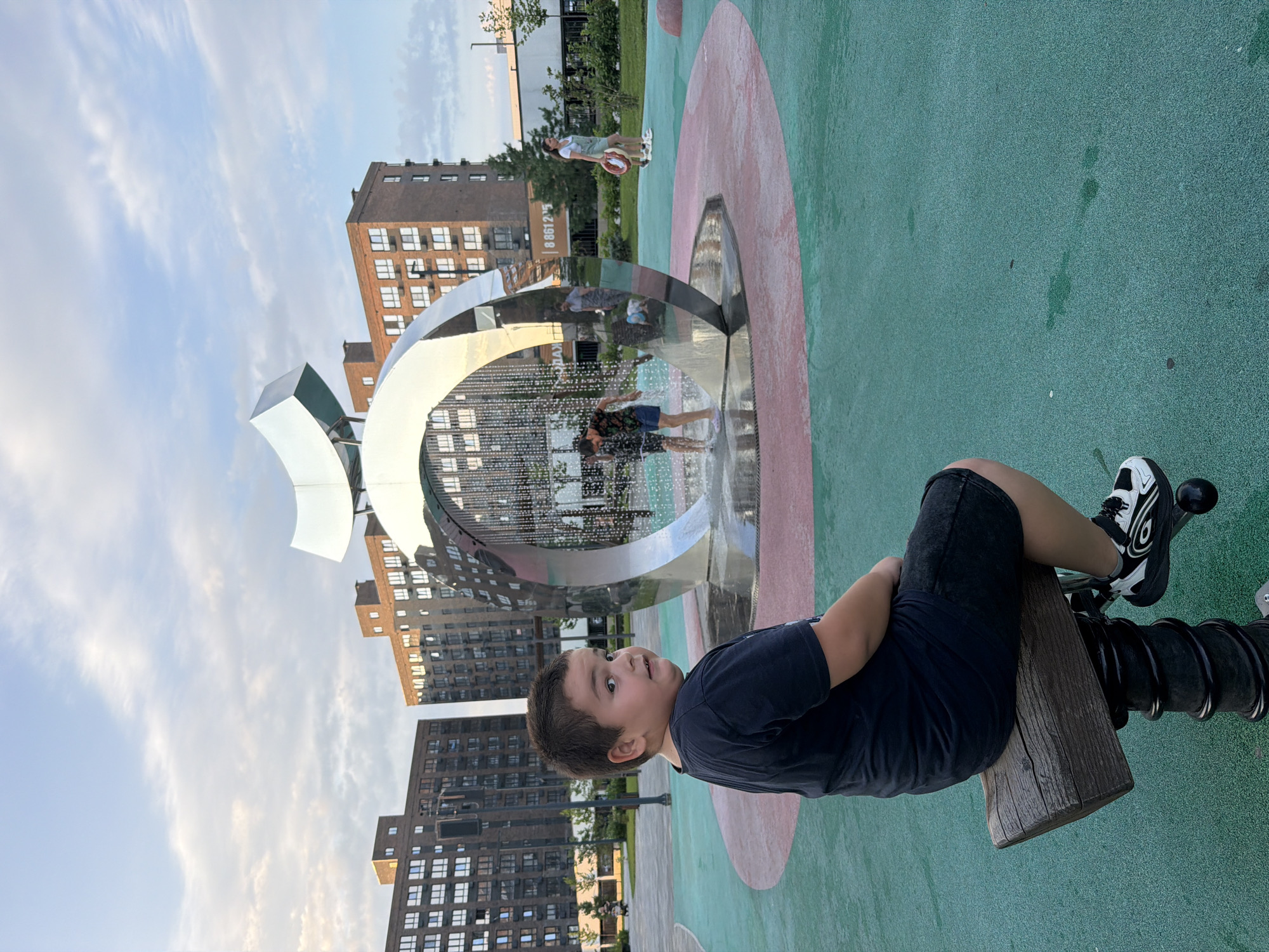 In an outdoor urban park, a young boy with dark hair is seated on a wooden seesaw, facing away from the camera. He is wearing a black t-shirt and black shorts. Behind him, a large, modern metal sculpture with water features is visible. In the background, several multi-story brick buildings with numerous windows line the horizon under a cloudy sky. Other people are present near the sculpture, some interacting with the water. The ground is a textured green surface with a red circular path.