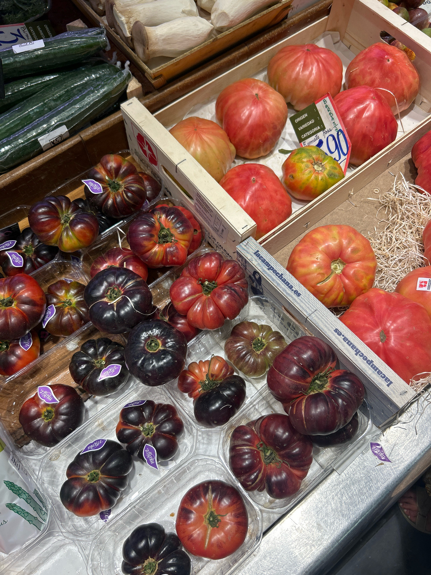 The image shows a close-up, overhead view of a display of heirloom tomatoes and cucumbers in a market stall, likely in Palma de Mallorca, Spain. The produce is arranged in wooden crates and clear plastic containers. Several varieties of tomatoes are visible, including large, ribbed red tomatoes, darker, almost black heirloom tomatoes with red streaks, and smaller, multi-colored heirloom tomatoes. Some tomatoes have purple stickers with the word "Mura" on them. Long, dark green cucumbers, wrapped in plastic, are stacked in a wooden crate on the left side of the image. In the background, there are some large white mushrooms in a wooden crate. The lighting suggests an indoor or covered market setting. No people are clearly visible, though the edge of a person's foot wearing sandals is visible at the bottom right. The overall impression is a fresh produce market stall showcasing a variety of high-quality, visually striking tomatoes and cucumbers.