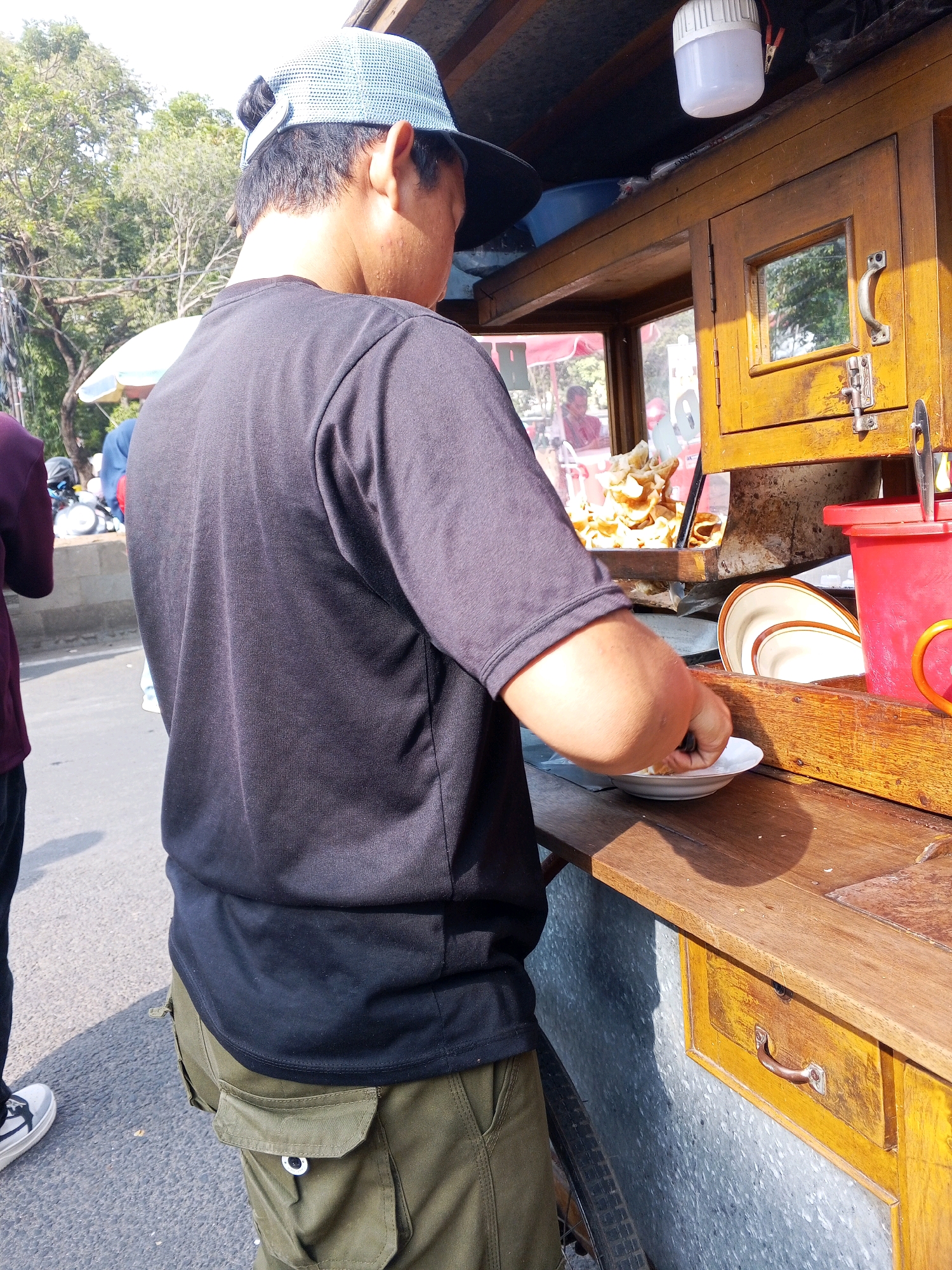 A man in a black t-shirt and green cargo pants stands at a wooden food cart on a street in Serang, Indonesia. The cart has a yellow wooden exterior with a glass display case filled with fried snacks. A red plastic container is visible on the right side of the cart. The man is reaching for a white plate, presumably to serve food. In the background, trees and other people are visible, suggesting a busy street scene. The lighting indicates it is daytime, with strong sunlight casting shadows.