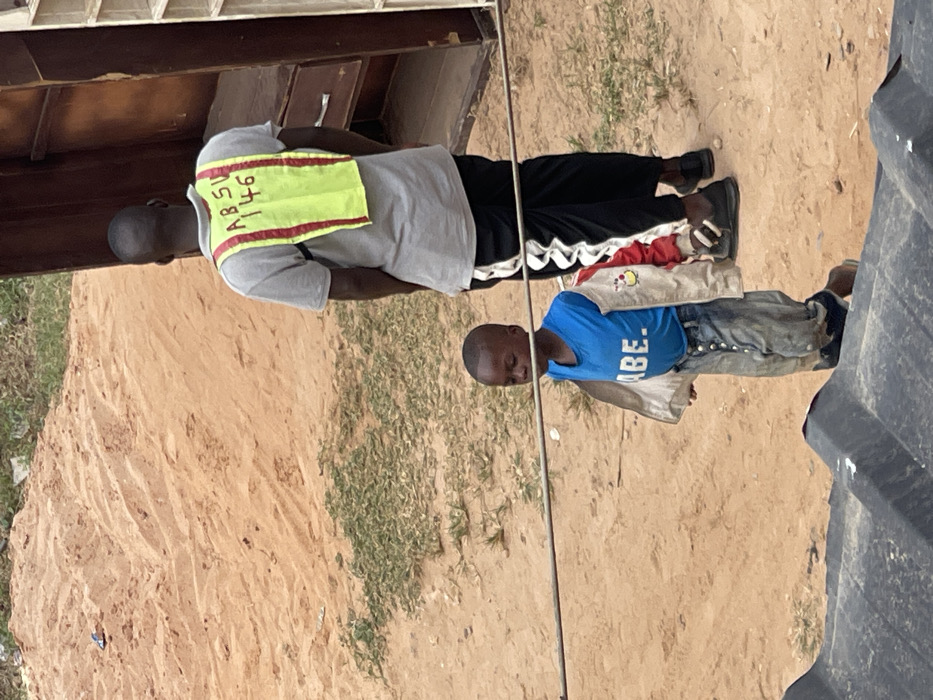 Two people stand in front of a sandy yard with sparse vegetation and a dark object on the right. The person on top wears a grey shirt, black pants with a white stripe, and a bright yellow vest with red trim and black writing. The younger person wears a blue shirt that reads "ABE," denim jeans, and a jacket. Both face the same direction. The top person is taller.