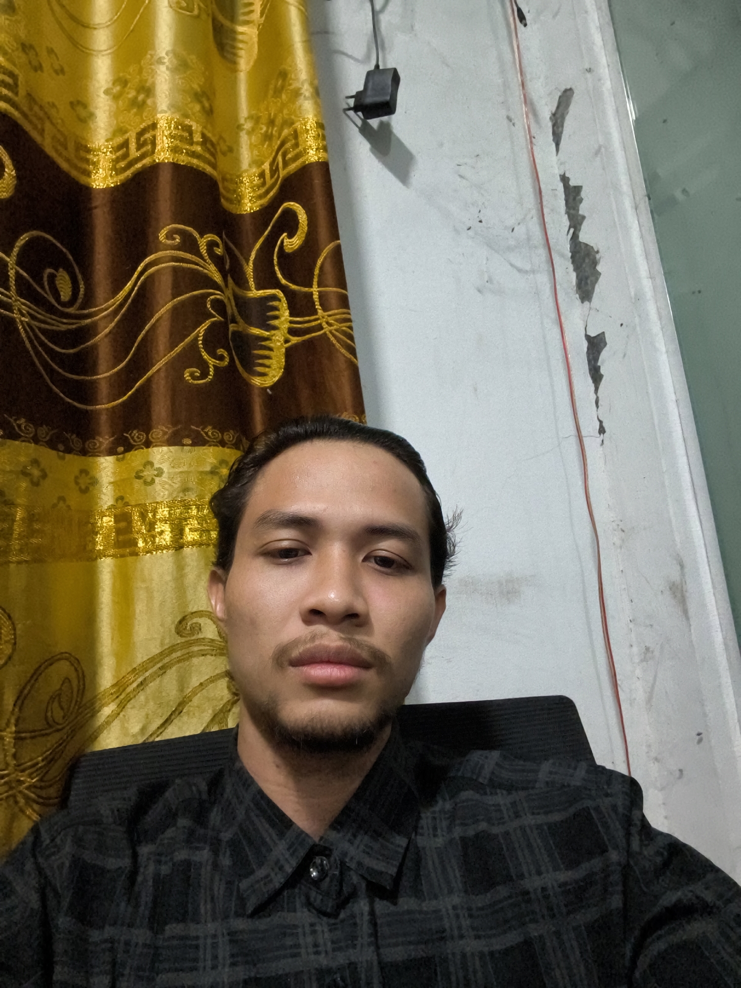 A medium shot shows a man with dark hair pulled back and a faint mustache, facing forward. He is wearing a black collared shirt with a subtle grey plaid pattern. The background features a brown and gold patterned curtain on the left, and a white wall with some peeling paint and a red wire on the right. A dark chair is visible behind the man. The setting appears to be an indoor residential area in Pulodolang, Indonesia.
