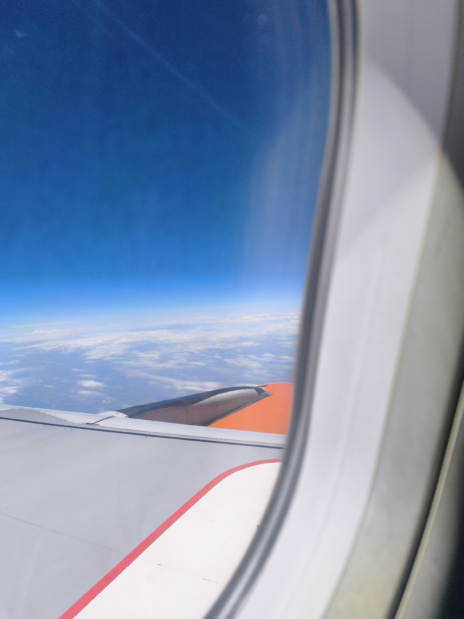 A vertical, full shot captures an aerial view through an airplane window. The deep blue sky occupies the top half of the frame, transitioning to a vast expanse of white, fluffy clouds below. The edge of an airplane wing, painted in gray with a thin red stripe, is visible in the foreground, extending from the bottom left corner. A portion of the wing's orange underside is also visible. The view is framed by the window's white interior structure. There are no people or discernable landmarks from the city of Trebbin, Germany, visible in this image.