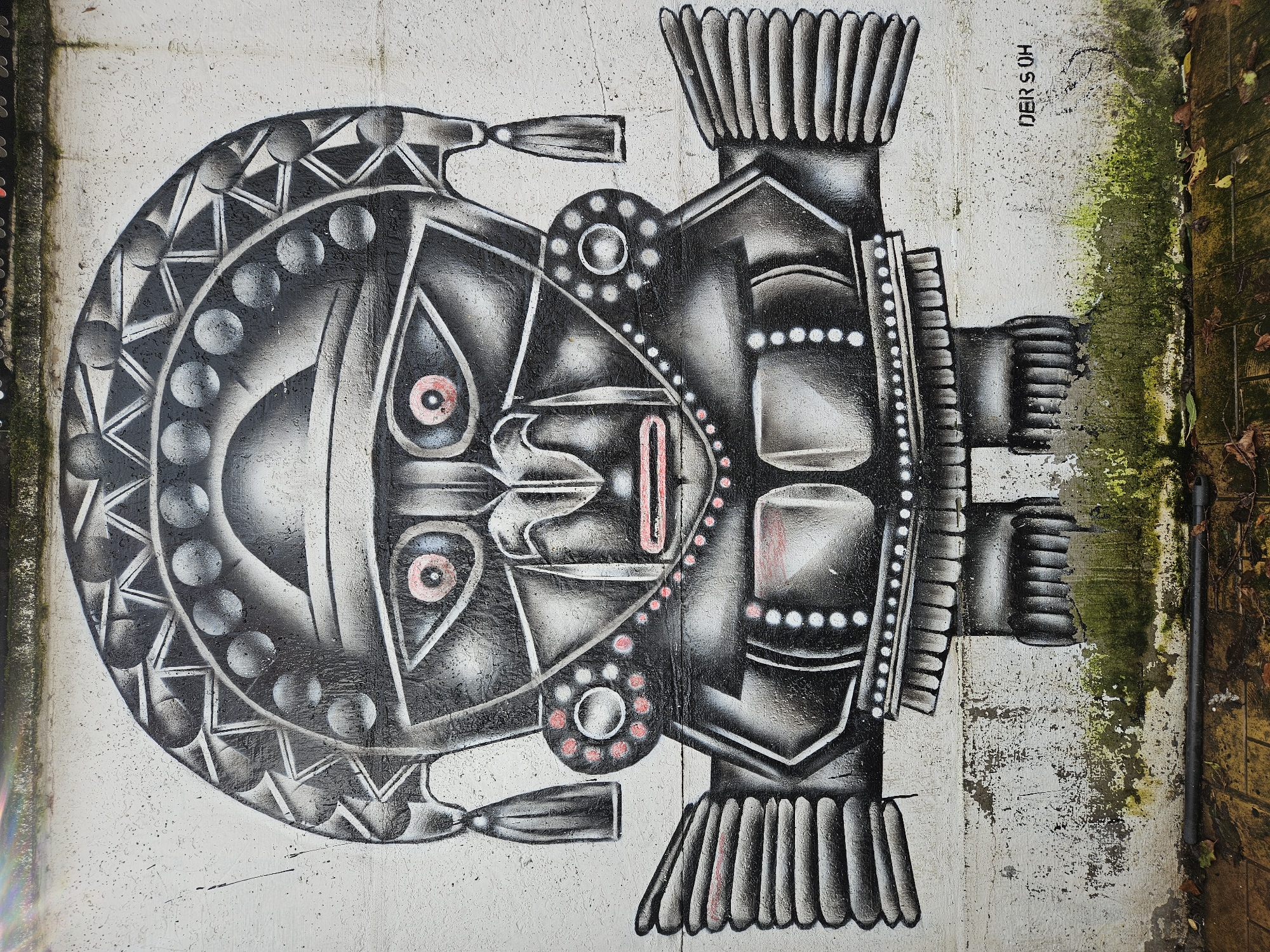A horizontally oriented eye-level shot depicts a piece of graffiti art resembling a stylized mask, possibly inspired by indigenous or tribal designs. The artwork is painted on a concrete wall, which shows signs of wear and weathering. Moss is visible on the wall's edges, suggesting damp conditions. To the far right, a portion of a brick wall can be seen.
The mask is rendered in shades of black, white, and gray, with small touches of red. The left side of the mask features a rounded shape with geometric patterns and circular motifs. The right side is more angular and symmetrical, with a series of rectangular forms and vertical lines.
In the top right corner of the image, a signature or tag is visible: "DER S OH." The scene appears to be outdoors, possibly in an urban or semi-urban setting. The location is in the city of Osorno, Chile.